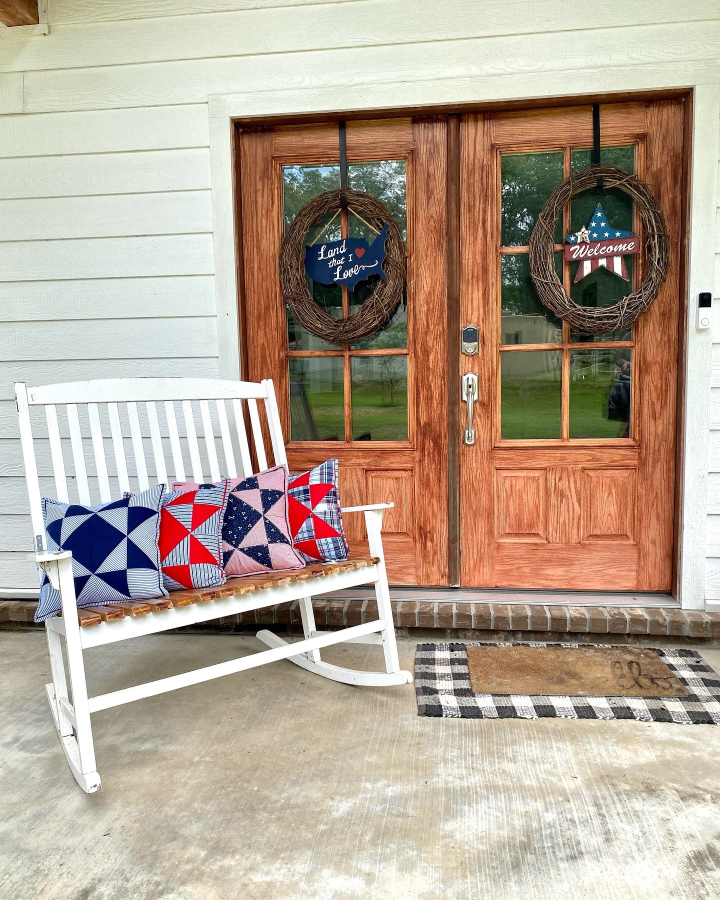 Decorative front door with two patriotic wreaths, a white rocking chair with patriotic pillows, and a welcome mat on the porch.