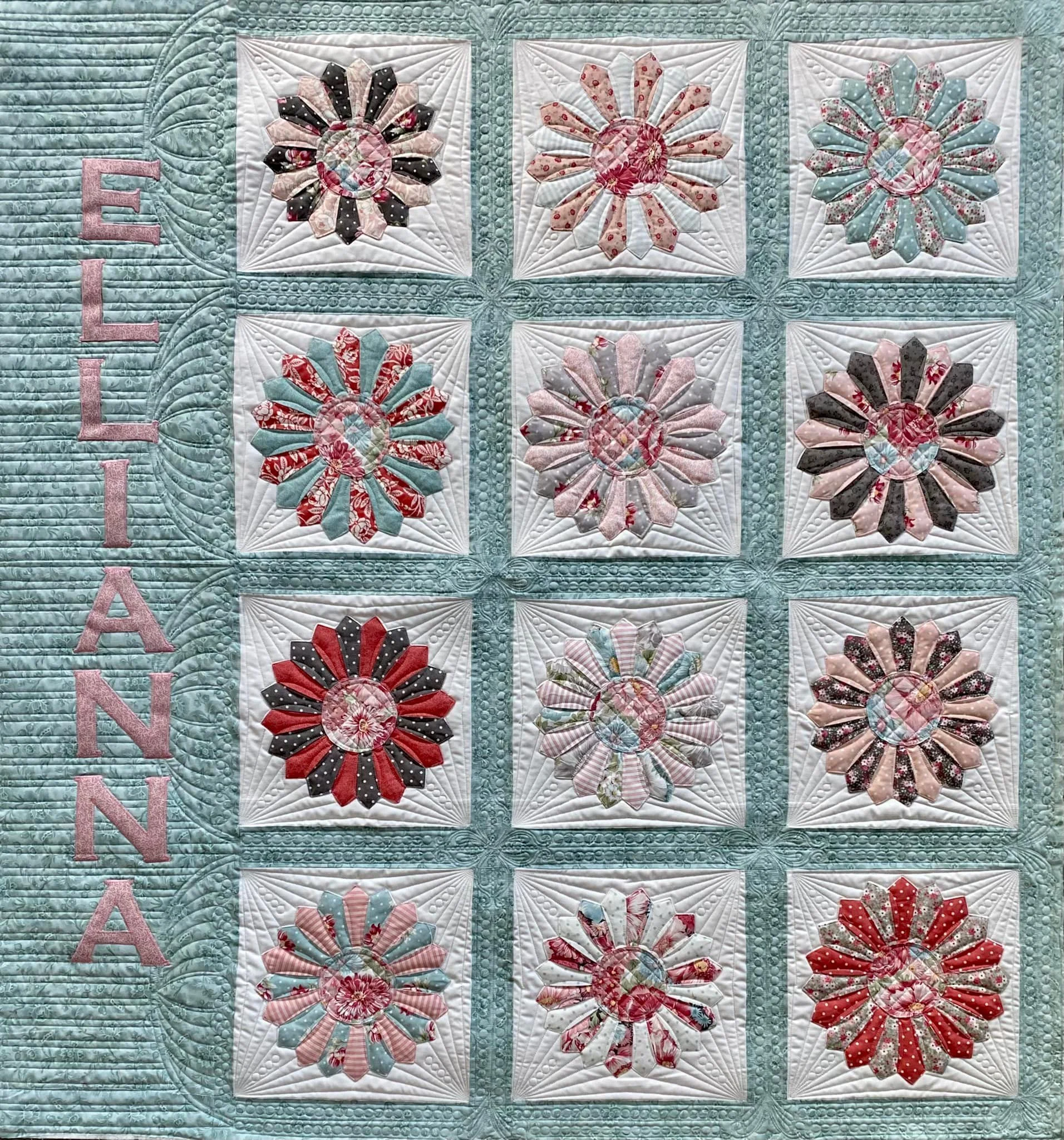 A quilt with a border of turquoise and pink letters spelling 'ELLIANNA' on the left side, and a grid of twelve fabric flower blocks arranged in three rows and four columns, each containing a circular floral pattern made of colorful petal shapes again