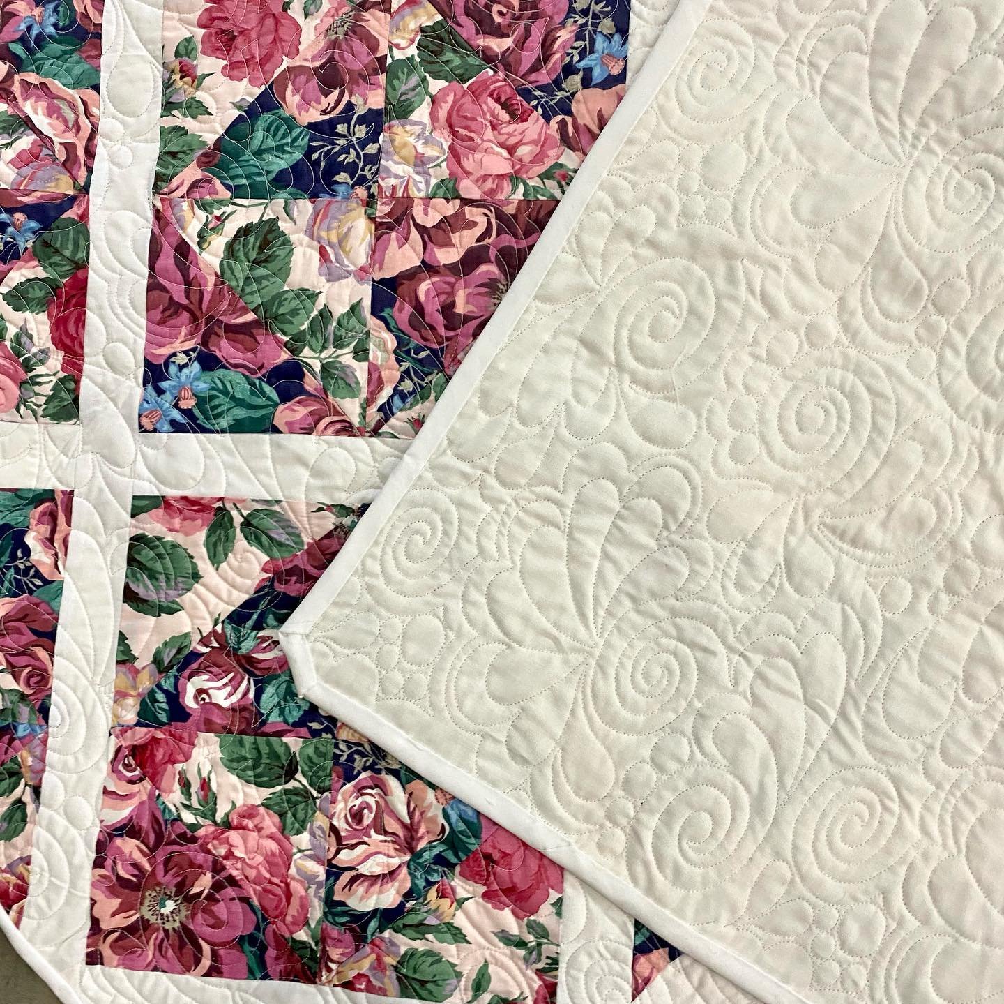 Close-up of a quilt with a floral pattern in shades of pink, red, green, and blue, with a white, decorated quilted backing with swirling and floral stitching patterns.