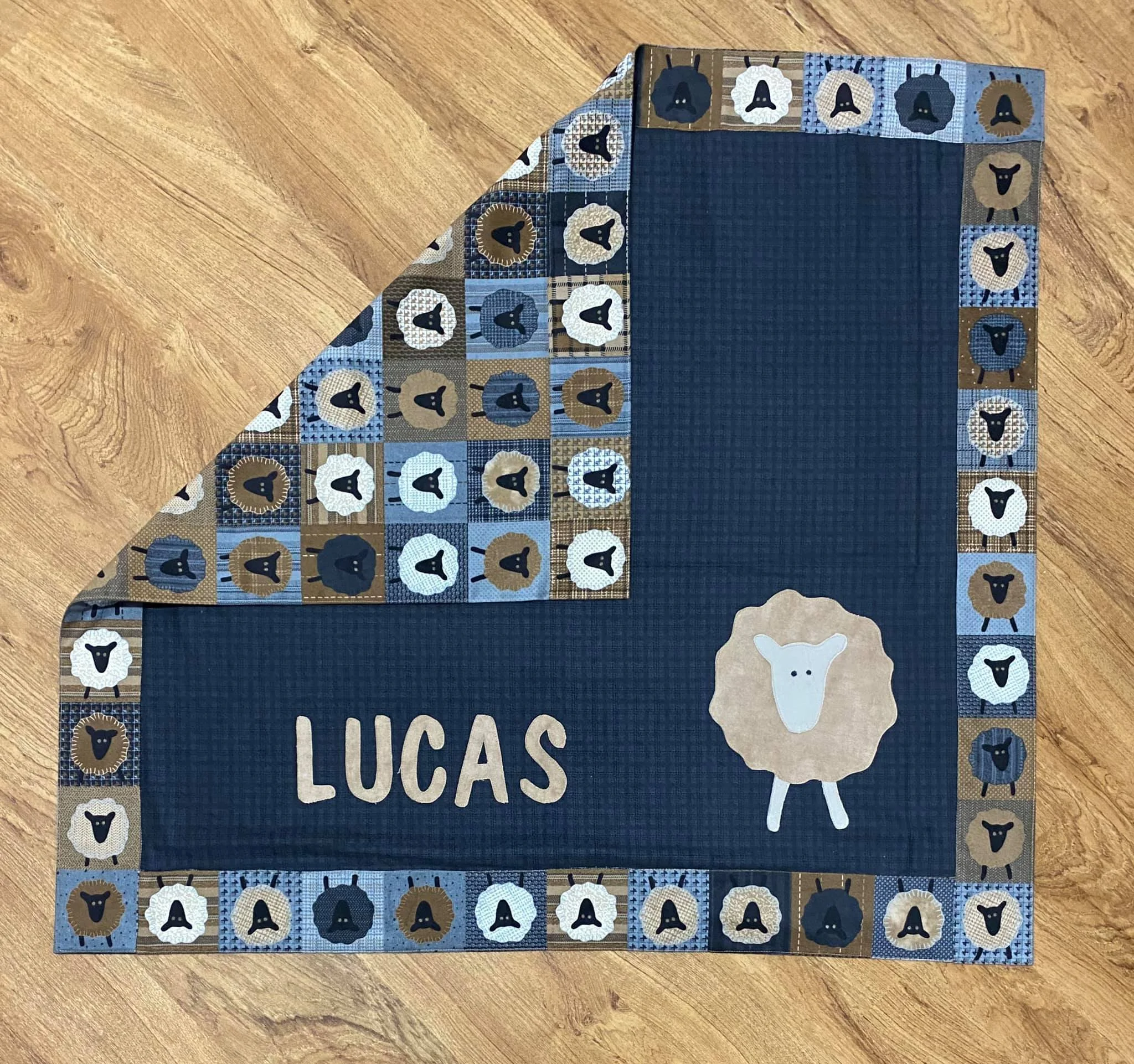 A personalized quilted blanket with a blue background, featuring the name "LUCAS" and a cartoon sheep face on it. The border has a pattern of small squares with sheep faces in black, white, and brown colors, on a blue, tan, and checkered background. The blanket is placed on a wooden floor.