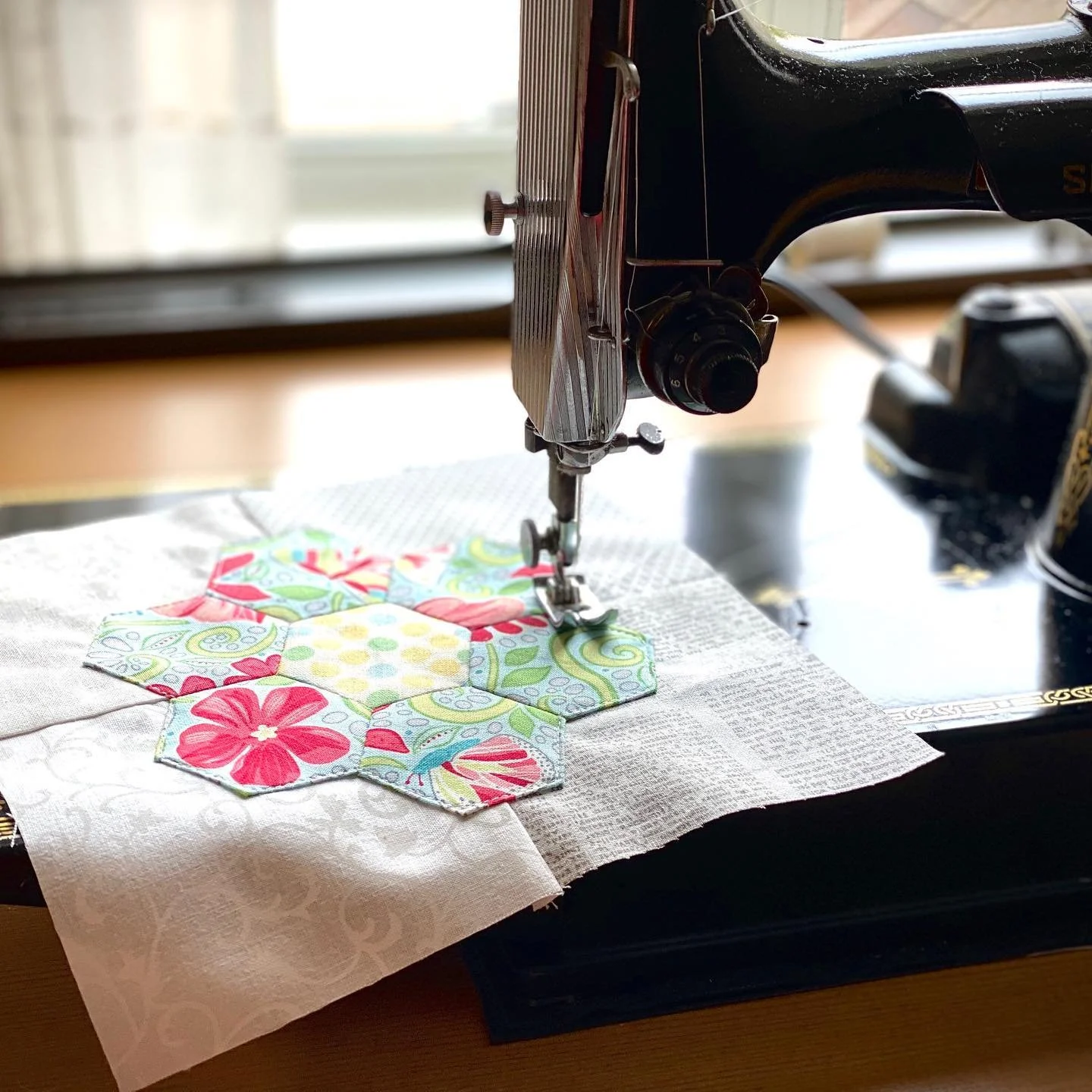 Close-up of a vintage sewing machine stitching a colorful quilt with floral and geometric patterns, placed on a table near a window.