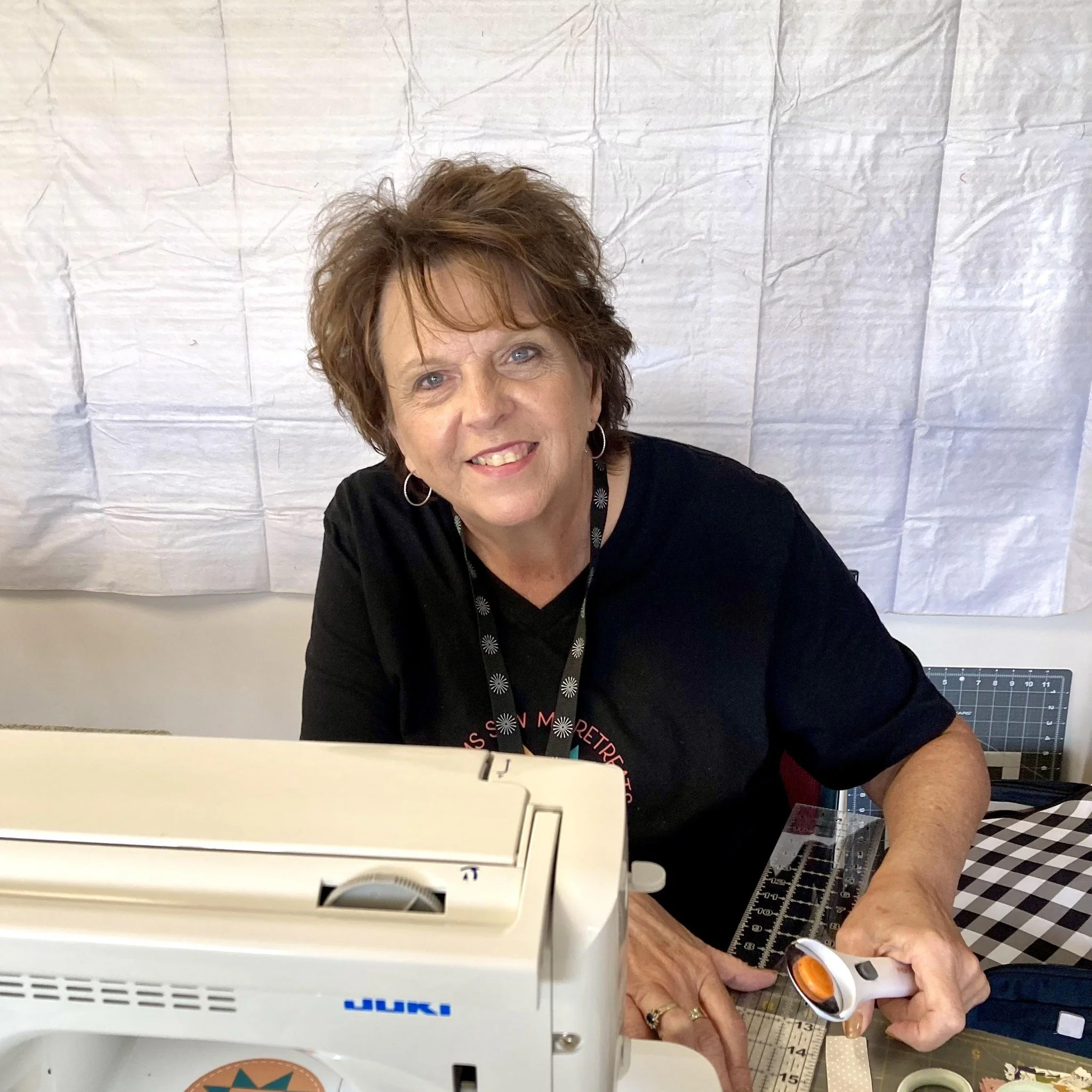 A woman with short brown hair, wearing a black shirt, sitting at a craft table with a sewing machine and a rotary cutter, smiling at the camera.