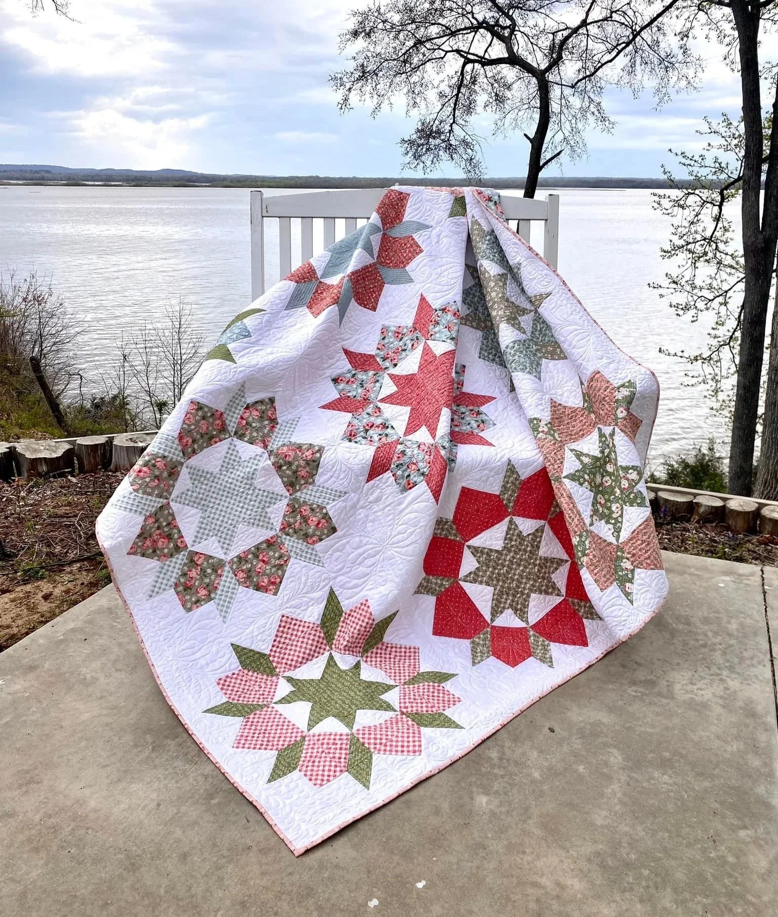 A quilt with holiday-themed star and poinsettia patterns draped over a white rocking chair near a river, with trees in the background.