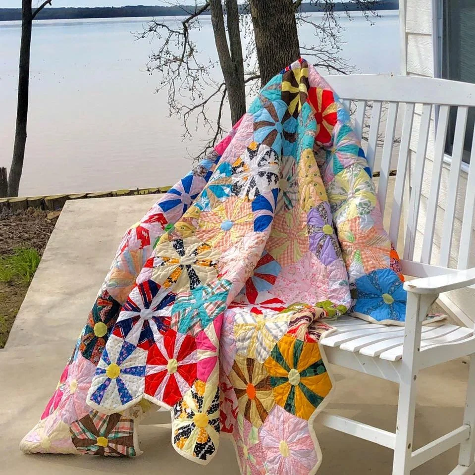 A colorful quilt with floral patterns draped over a white outdoor rocking chair on a porch overlooking a river with trees.