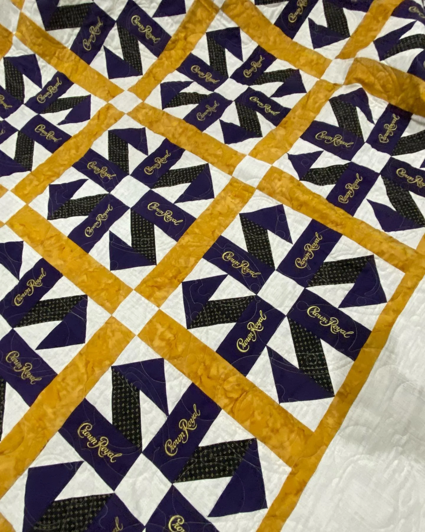 A quilt with a blue, black, and white star pattern and yellow-orange borders, featuring the words 'Crown Royal' in gold embroidered on some of the squares.
