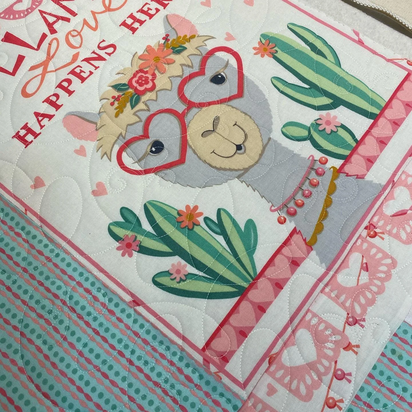 Close-up of a colorful fabric with a cartoon llama wearing heart-shaped glasses, surrounded by cacti, flowers, and hearts, with a partially visible pink border and text.