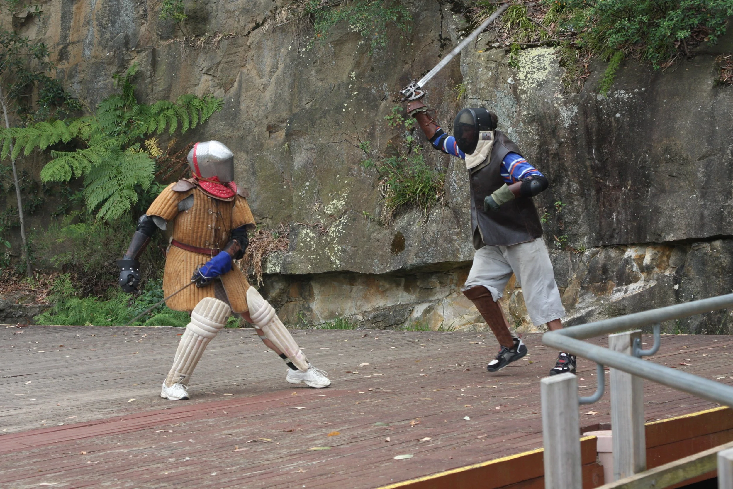 Two individuals engaged in a medieval-style sword fight on a wooden platform, with one person holding a sword in an attacking position and the other person in a defensive stance. Both are dressed in medieval armor and protective gear, including helme