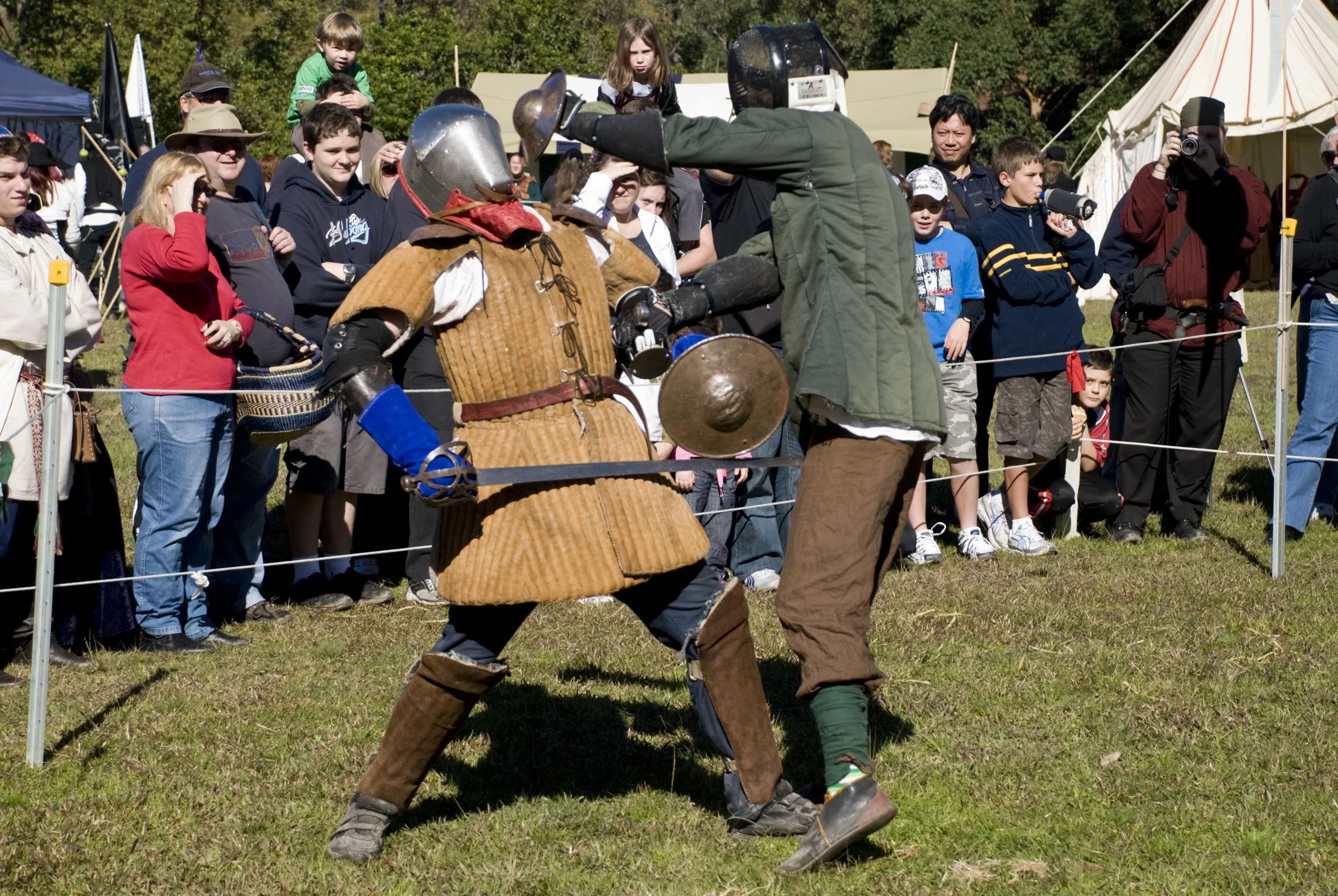 Two people dressed in medieval armor engaged in a mock sword fight during a reenactment at a fair, with a crowd of onlookers watching behind a small barrier on a grassy field.