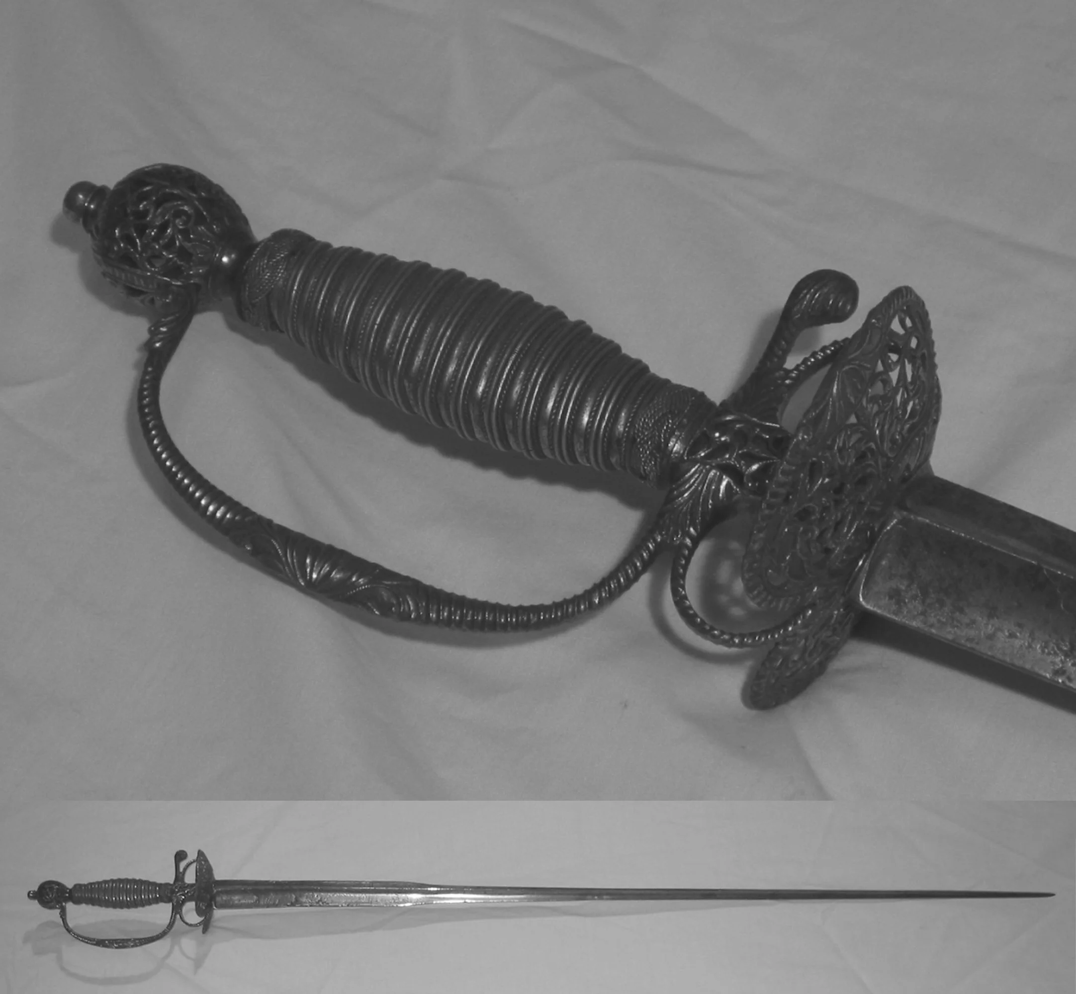 Black and white photograph of an ornate decorative sword with a detailed handle and guard, and a straight, slender blade, resting on a fabric surface.