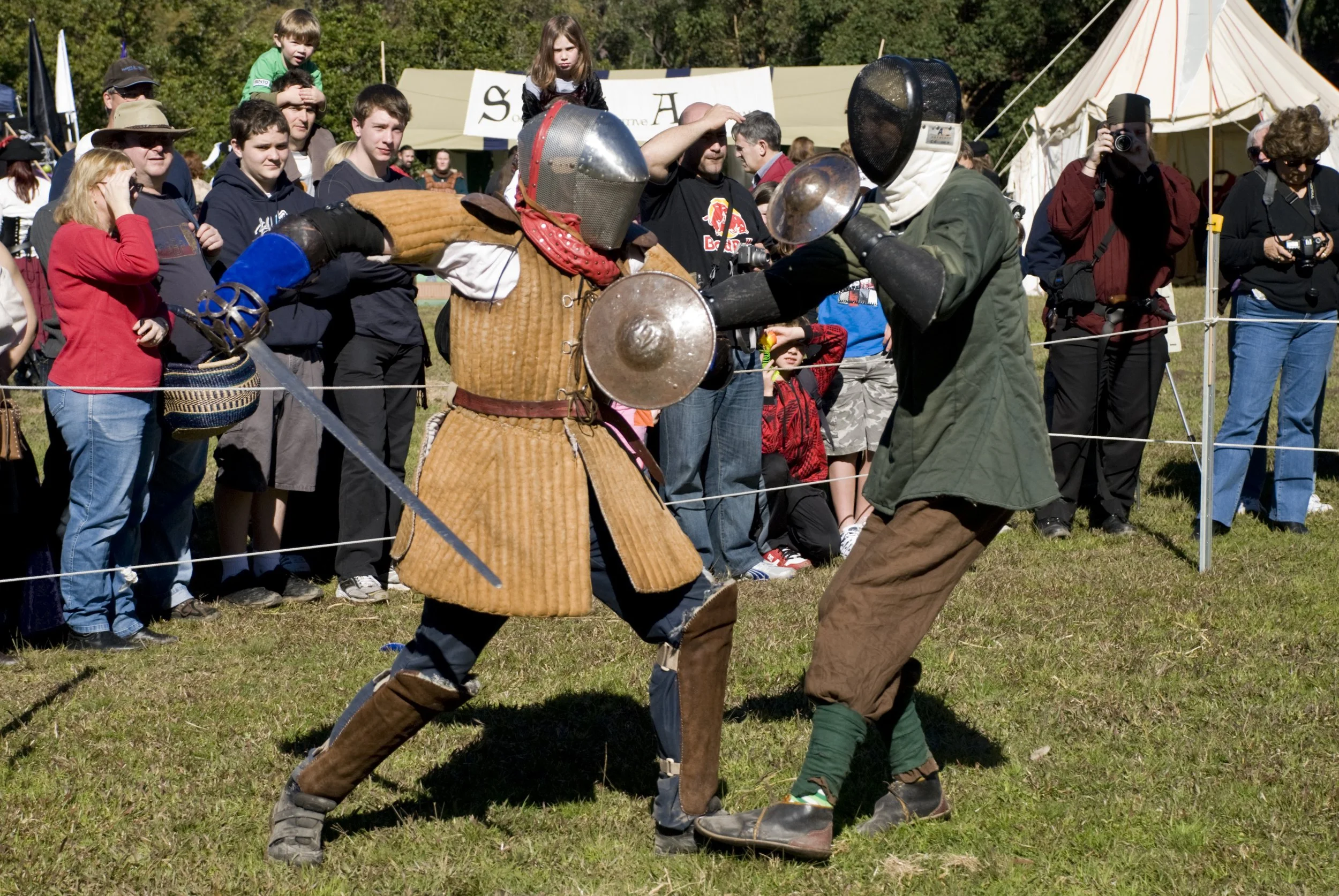 Two people in medieval costumes sparring with swords and shields while surrounded by spectators at a Renaissance fair.