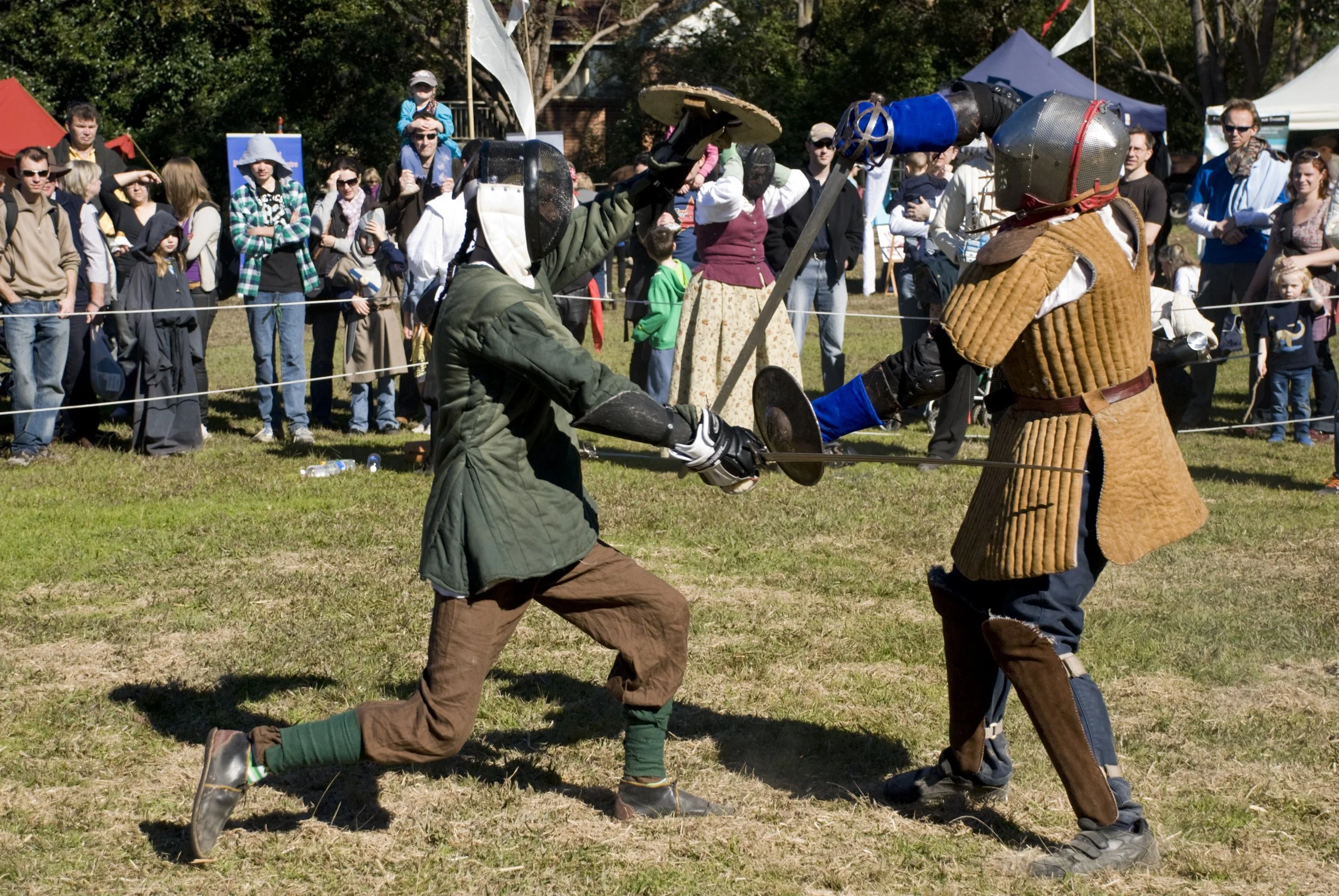 Two people dressed in medieval armor and clothing engaging in a sword fight at a Renaissance fair, with a crowd of spectators watching in the background.