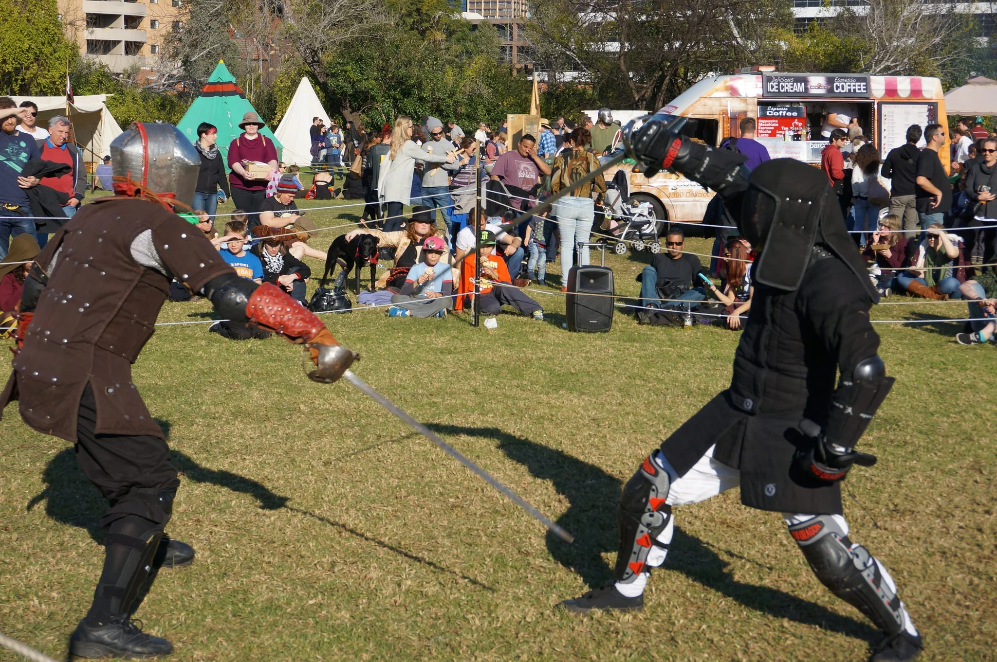 Two people in medieval armor practicing fencing on a grassy field at an outdoor event, with a crowd of spectators and tents in the background.