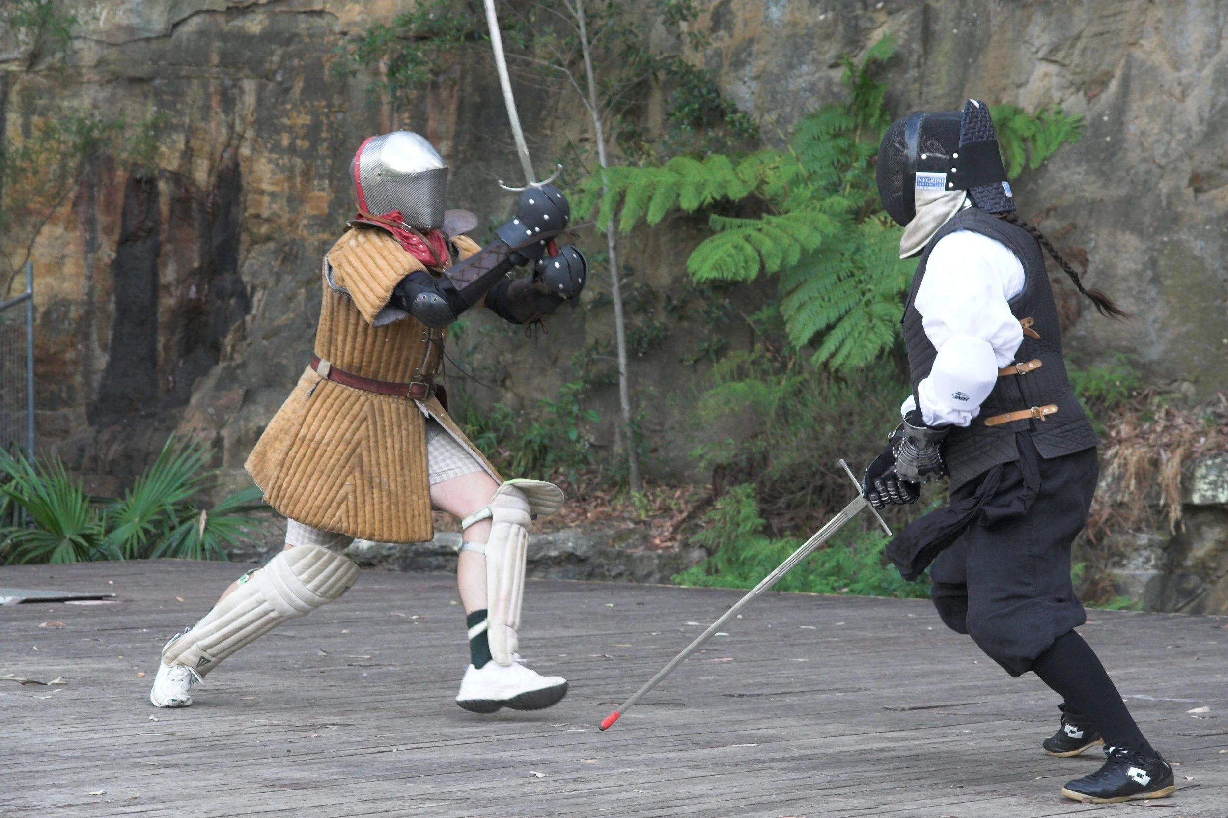 Two individuals engaged in a fencing match dressed in historical armor and clothing, outdoors on a wooden surface with a rocky and leafy background.
