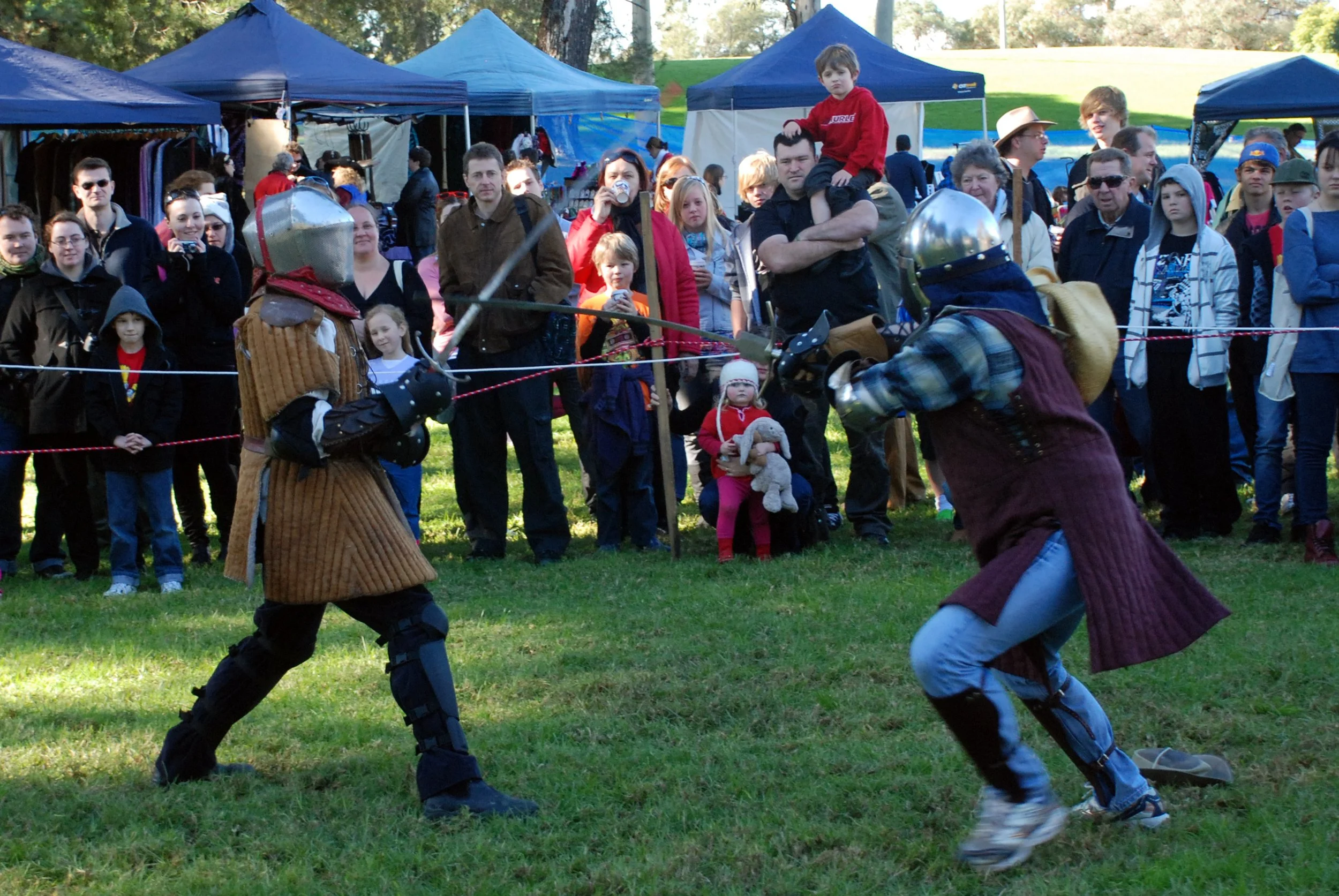 Two people in medieval armor and clothing engaged in a friendly sword fight at an outdoor event, with a large crowd watching.