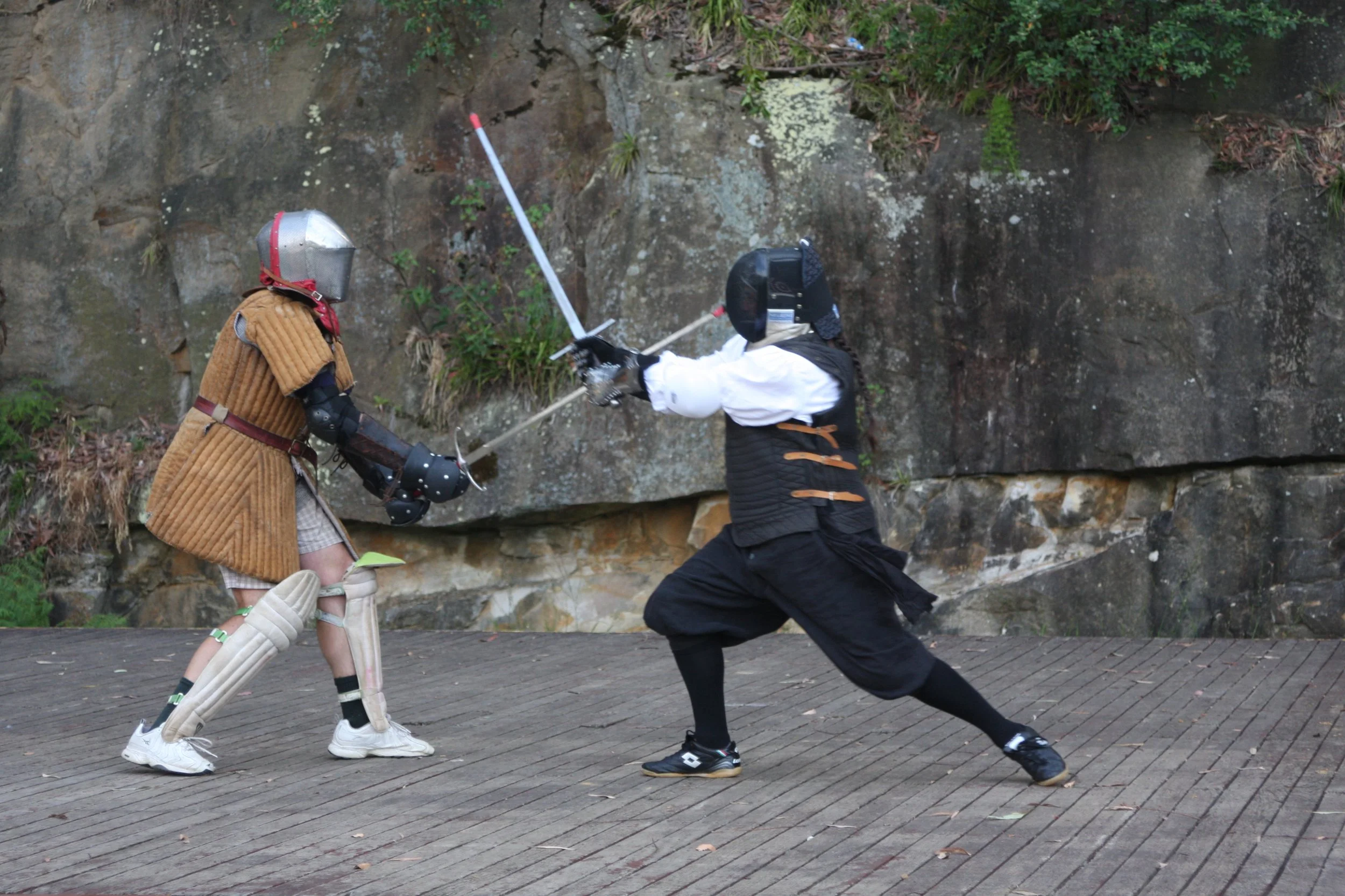 Two people dressed as medieval knights engaging in a mock sword fight outdoors on a wooden platform, with a rocky wall and greenery in the background.