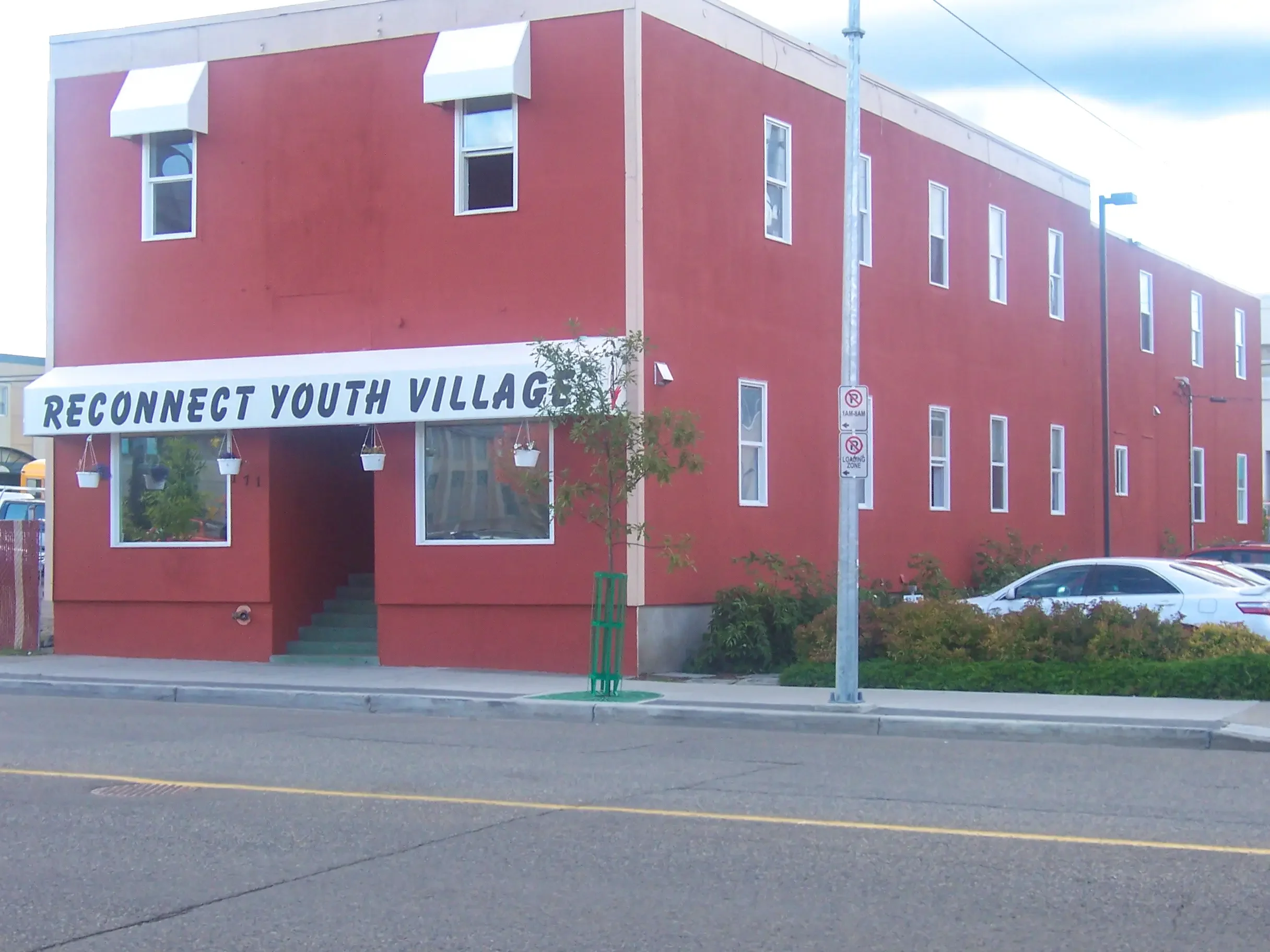 A red building with a white sign that says 'Reconnect Youth Village,' three hanging flower pots, and multiple windows. There are cars parked on the street and sidewalk with a small tree and bushes.