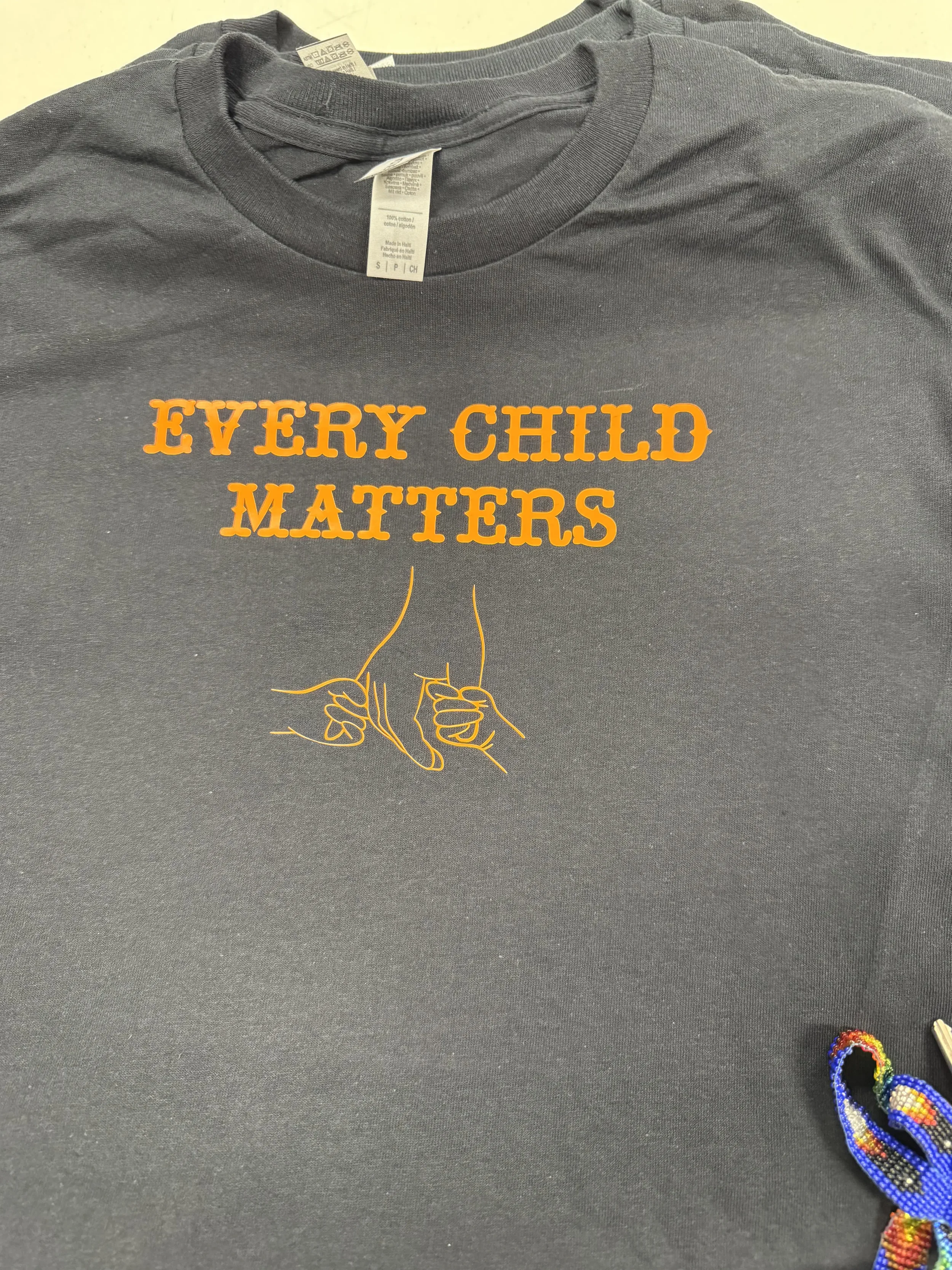 Black t-shirt with orange text that reads 'Every Child Matters' and an orange line drawing of two hands intertwined.