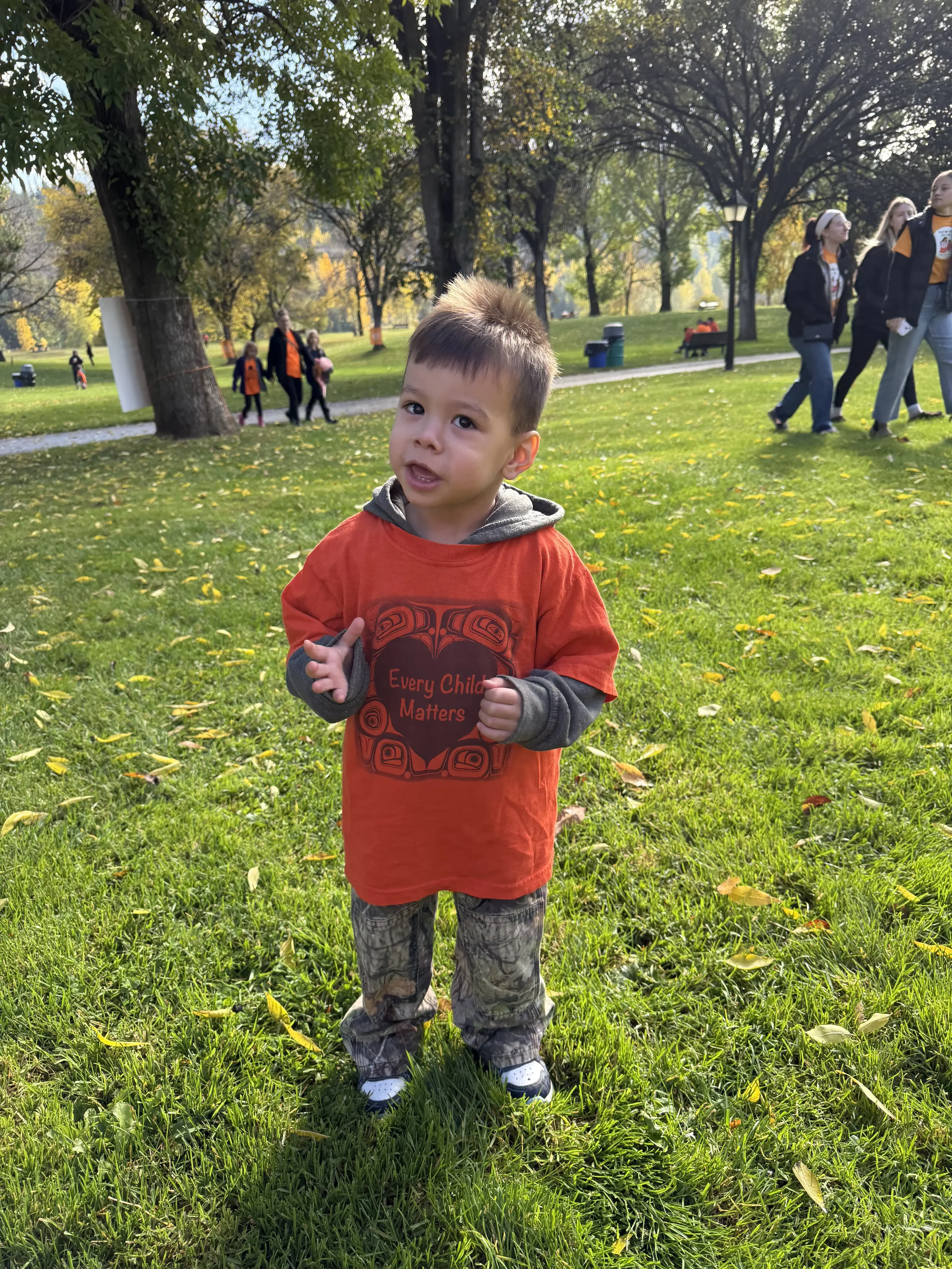 Young boy standing on grass in a park during fall, wearing a red shirt with a message, gray hoodie, and camo pants, with trees and other people in the background.