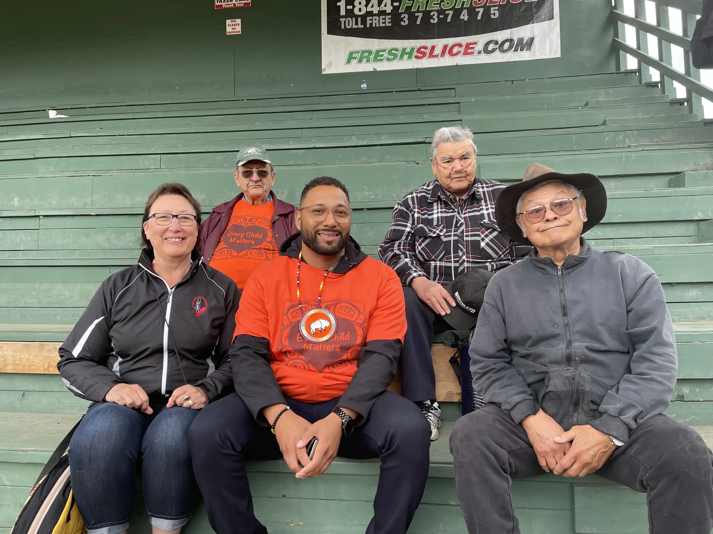 Five people sitting on green bleachers at an outdoor event, with a large banner behind them advertising FRESHSLICE.com and a phone number.
