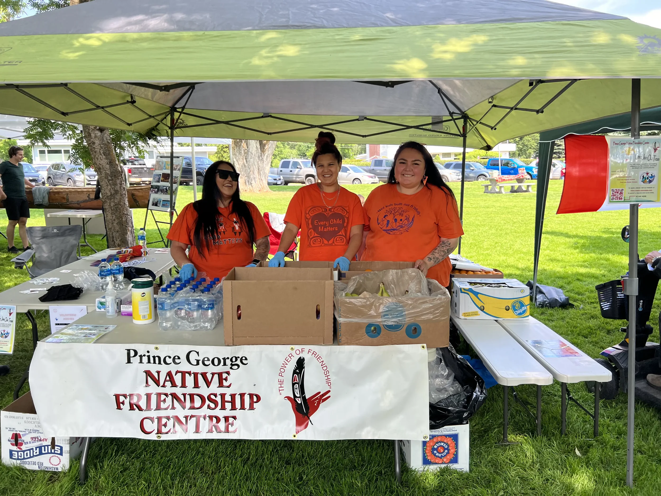 Three women smiling behind a table at an outdoor event, representing Prince George Native Friendship Centre, with supplies, bottled water, flyers, and boxes on the table, under a green canopy.