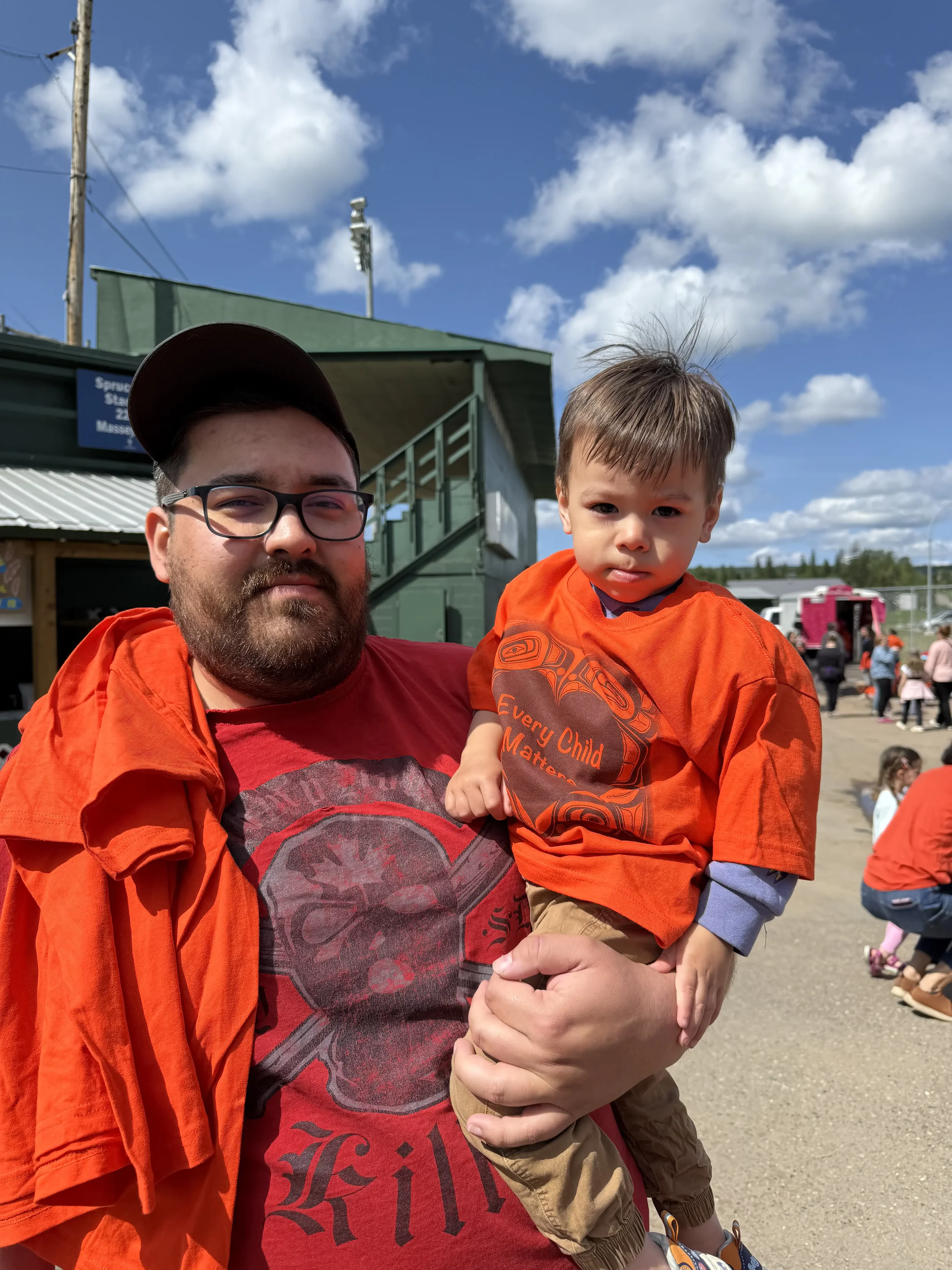 A man with glasses and a beard holding a young boy in an outdoor setting on a bright, sunny day. The man is wearing a red T-shirt with a graphic design and has an orange jacket draped over his shoulders; the boy is wearing an orange T-shirt and khaki