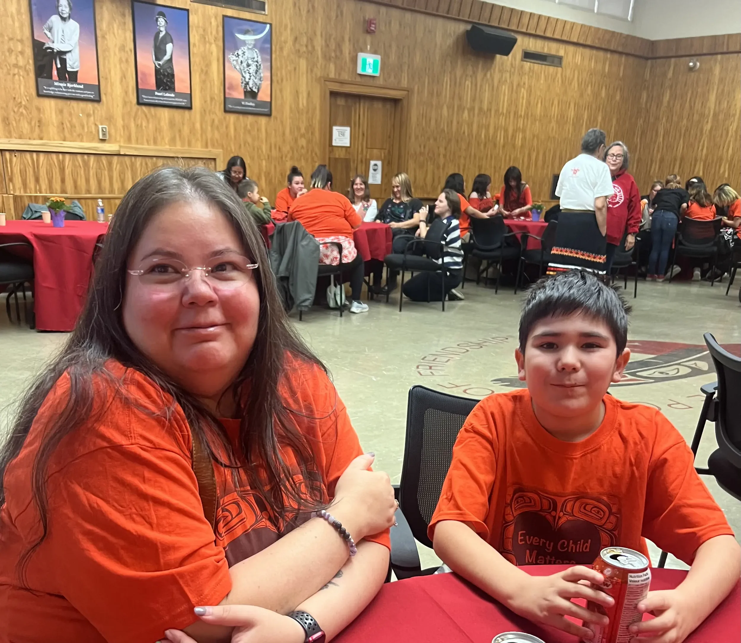 Two children sitting at a table during a gathering, with multiple people in the background at tables and a wood-paneled wall with portraits.