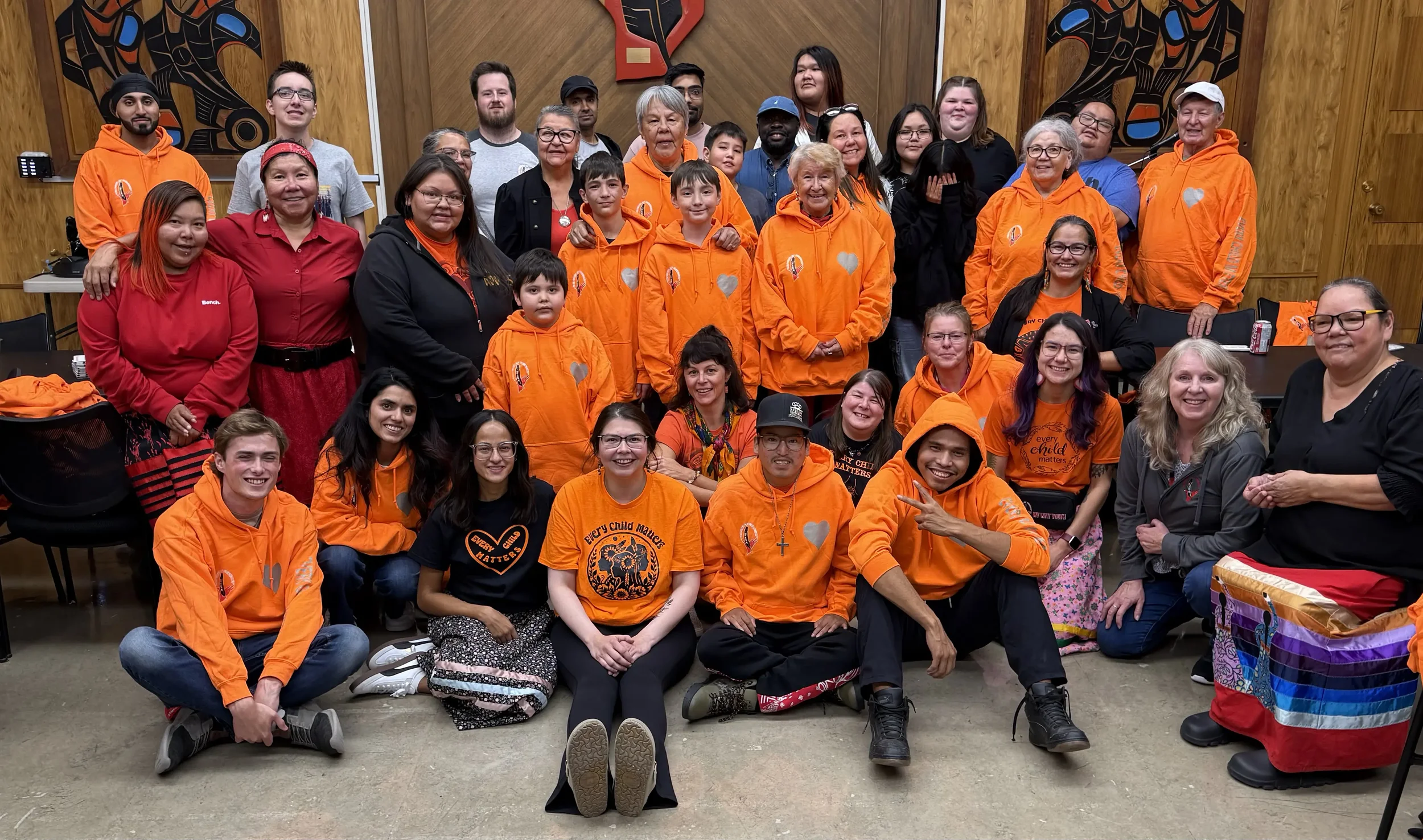 A large group of people, diverse in age and ethnicity, gathered indoors for a group photo. Many are wearing bright orange shirts and hoodies with a heart and message about every child matters. The background features wooden walls with indigenous artw