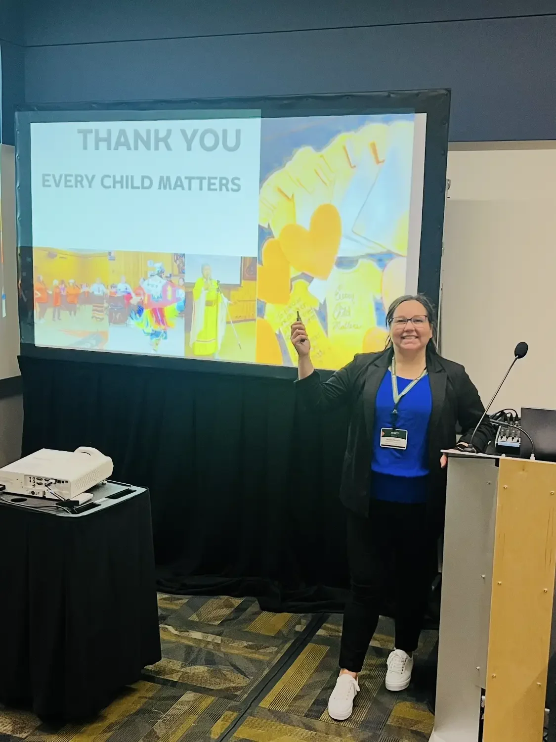 A woman standing in front of a presentation screen, smiling while holding a laser pointer. The screen displays a thank you message with the words "THANK YOU" and "EVERY CHILD MATTERS" along with colorful images of children, balloons, and balloons sha