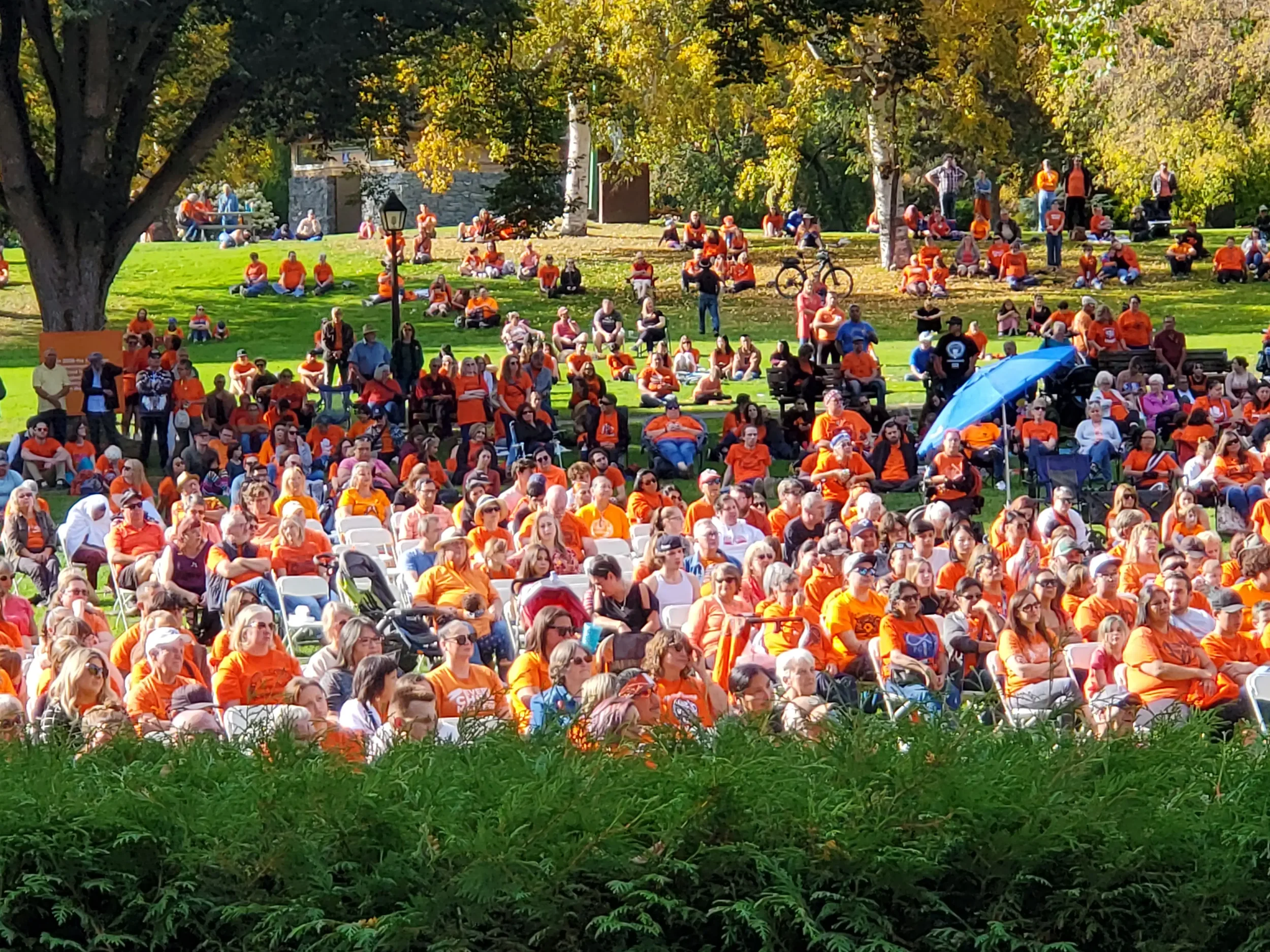 Large outdoor gathering in a park with many people wearing orange shirts, sitting on chairs and on the grass, some standing, with trees and a clear day in the background.