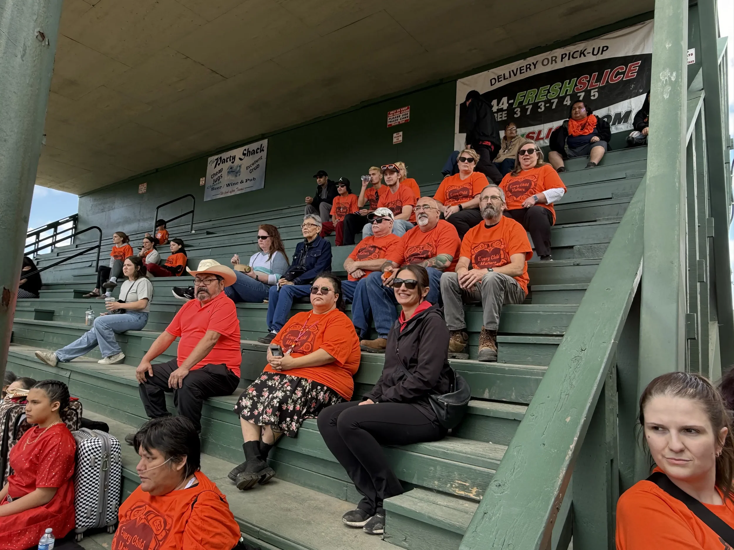 A group of people sitting on green bleacher seats at an outdoor stadium, some wearing orange shirts, with advertisements visible in the background.