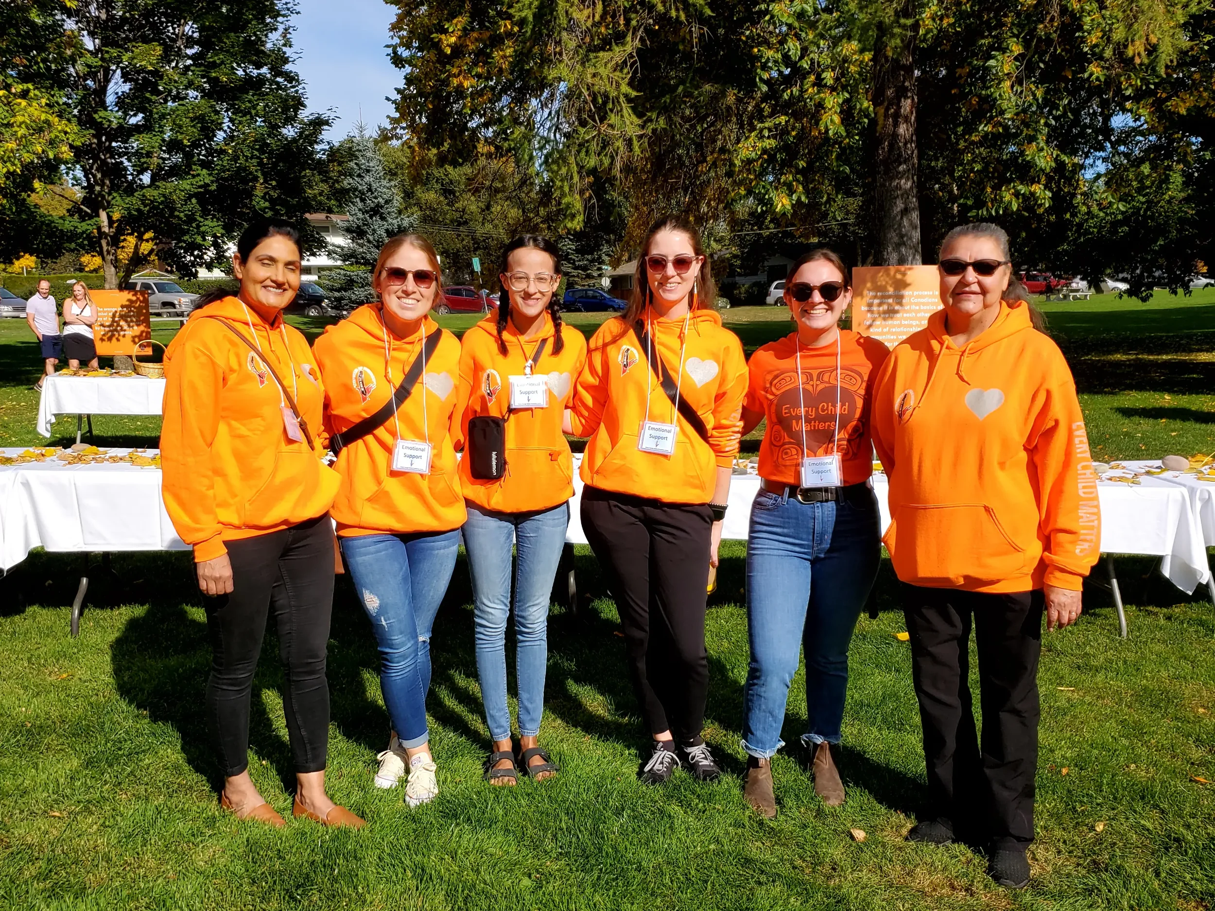 Group of six women standing outdoors on a grassy area, all wearing orange hoodies with white heart logos, at a community event or gathering.
