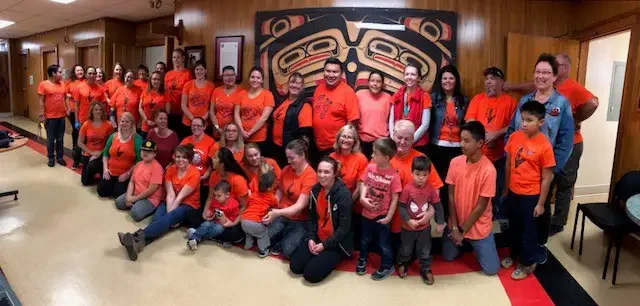 Group of children and adults posing together indoors, most wearing orange shirts, in front of a Native American artwork.