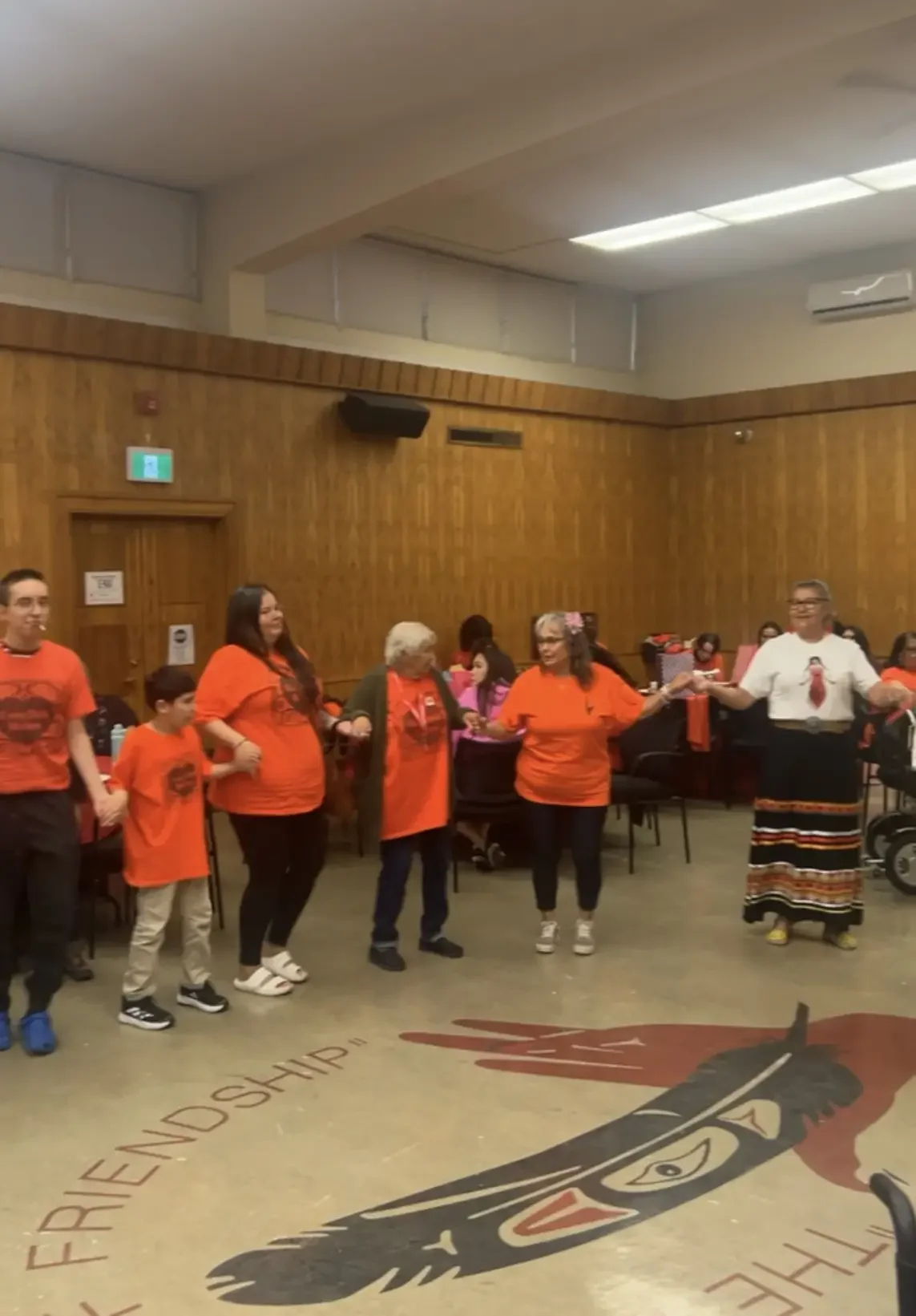 Group of people holding hands inside a community hall, wearing orange shirts, with some children and elderly women, and a person in traditional attire.