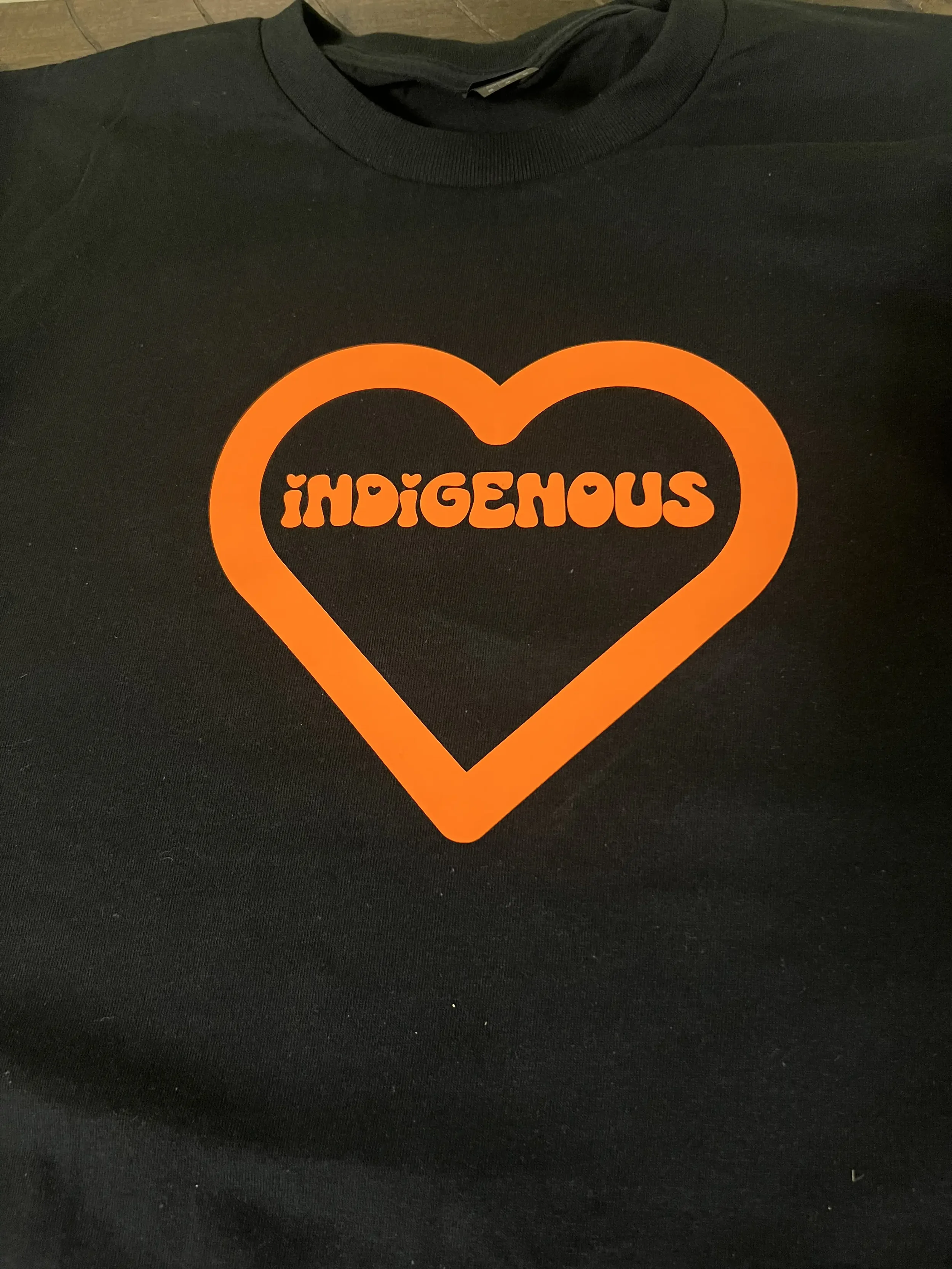 Black T-shirt with an orange heart outline and the word 'indigenous' written inside in orange with small heart accents in the letters.