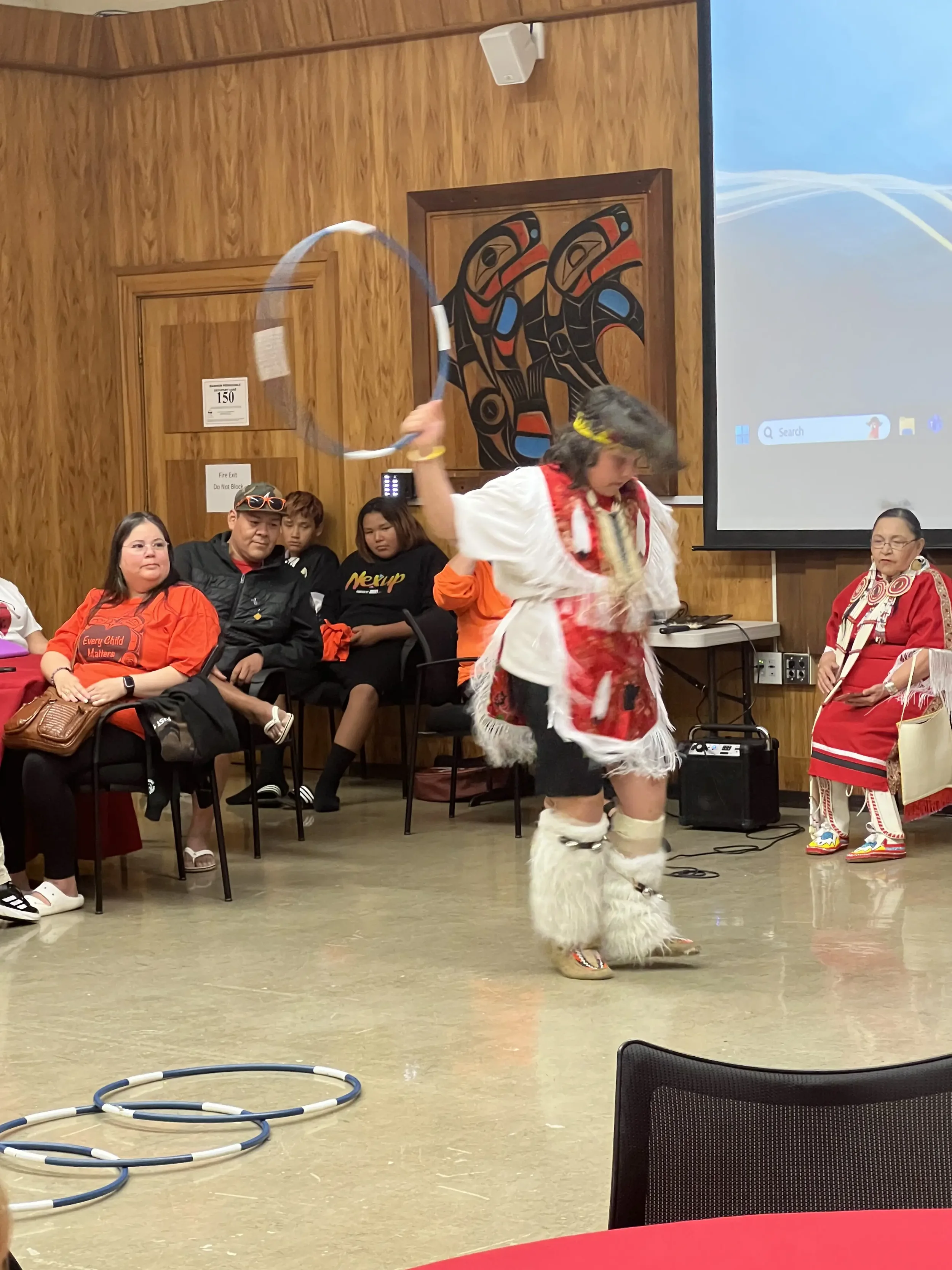 A woman dressed in traditional Indigenous attire, including furry leg covers and a red and white beaded dress, is spinning a hula hoop inside a room with wooden paneling. Several seated people in casual clothing are watching her perform. There is a l