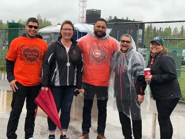 Group of five people standing outdoors in rainy weather, some wearing rain ponchos, smiling at the camera, with a chain-link fence, trees, and cloudy sky in the background.