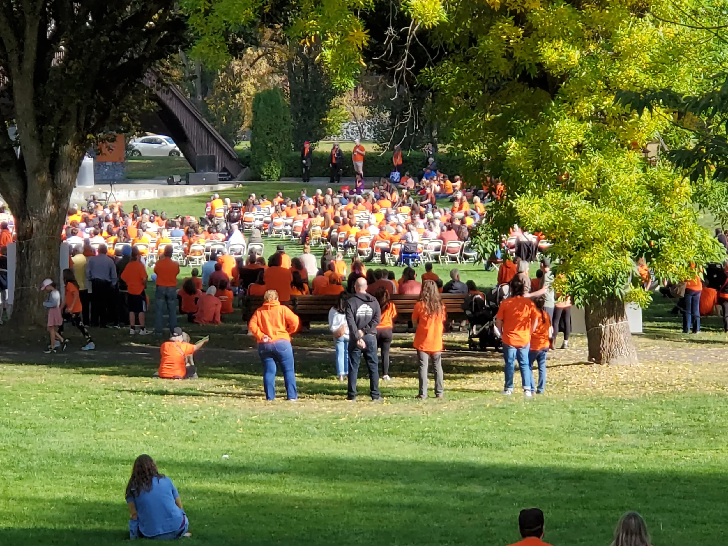 A large crowd gathered in a park, many wearing orange shirts, sitting on chairs and standing on grass, watching a performance or event on a stage under trees on a sunny day.