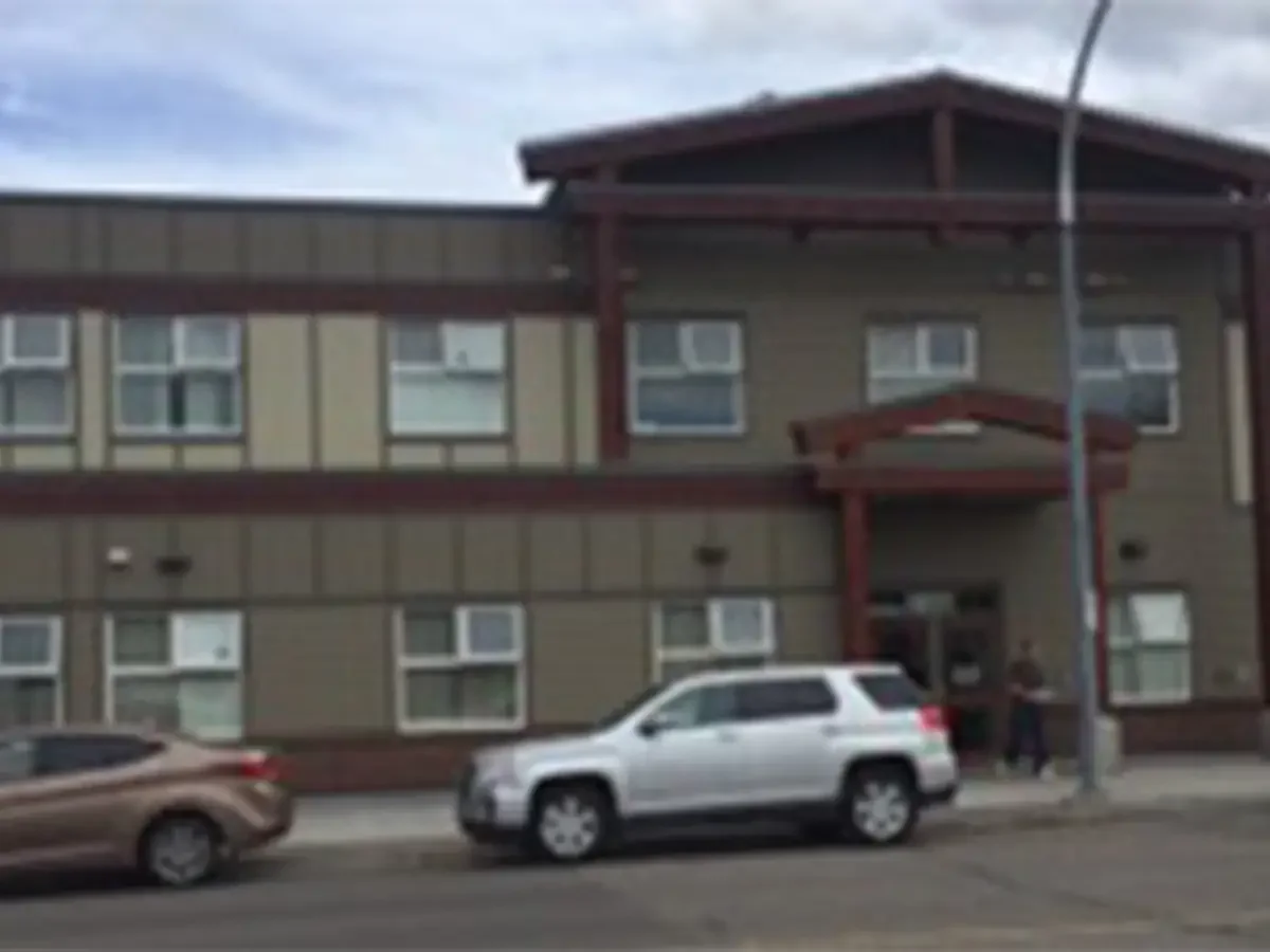 A multi-story apartment building with beige siding and red trim, several windows with Air conditioning units, a small porch at the entrance, a gray SUV parked on the street, and a brown car nearby, with two people standing at the entrance and a streetlamp in front.