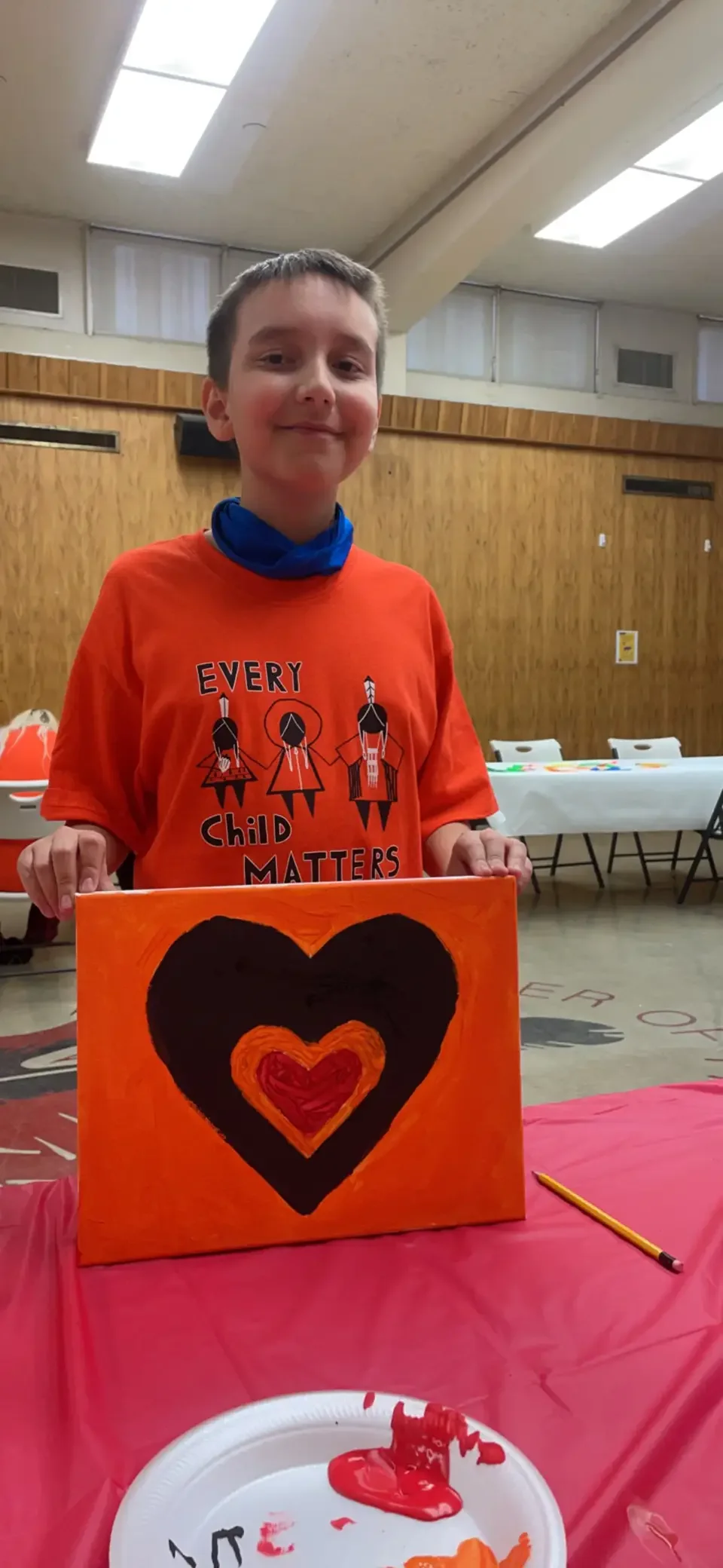 A young boy wearing an orange shirt with the words "Every Child Matters" and illustrations of three figures. He stands behind a table with a red tablecloth, holding a painting of a large black heart with an orange and red heart inside it. There are a
