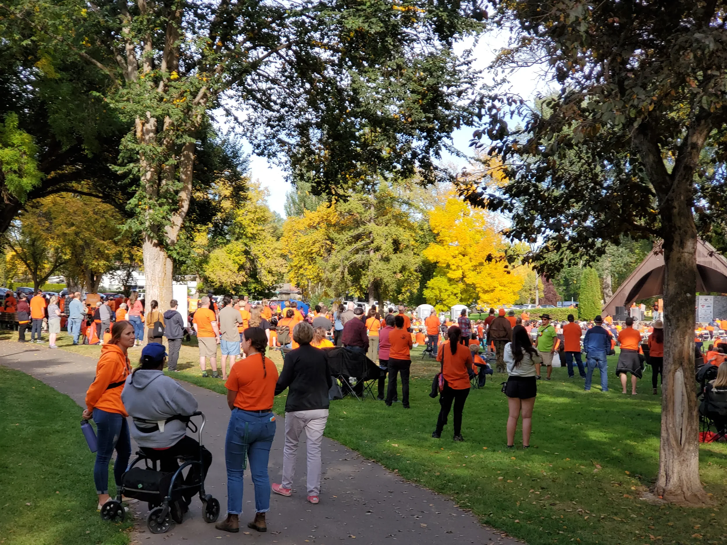 Crowd gathered in a park with trees, some wearing orange clothing, possibly for a gathering or parade.