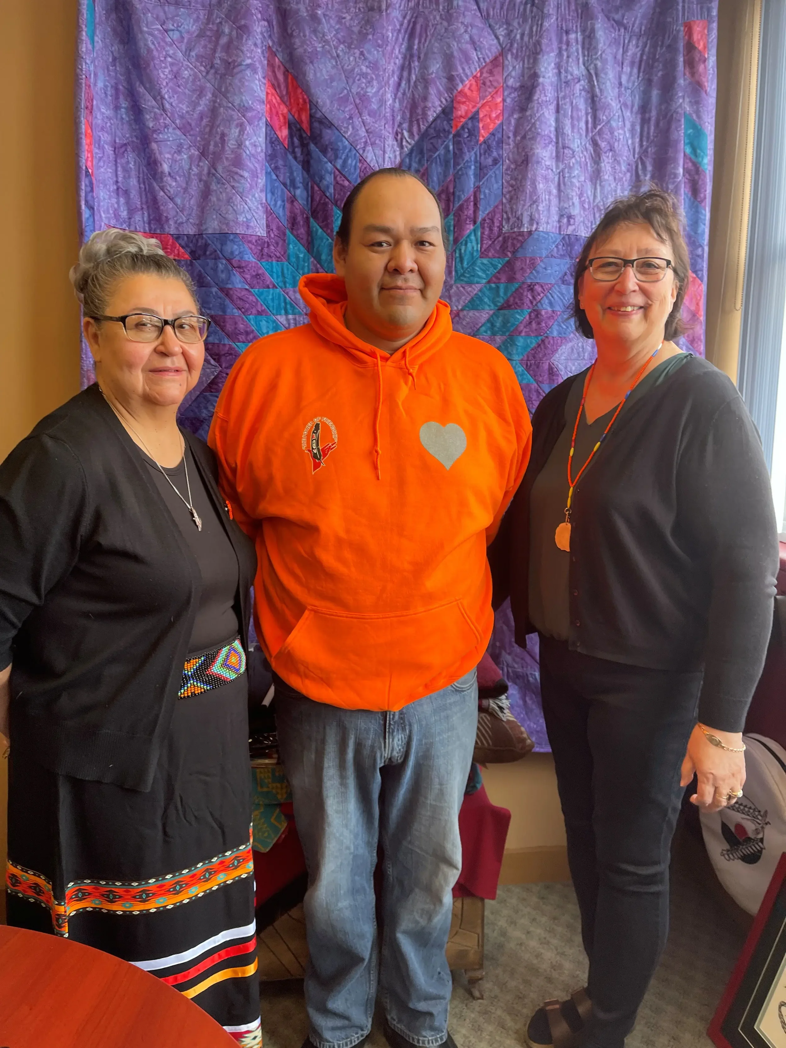 Three people standing indoors in front of a colorful quilted wall hanging, smiling at the camera.