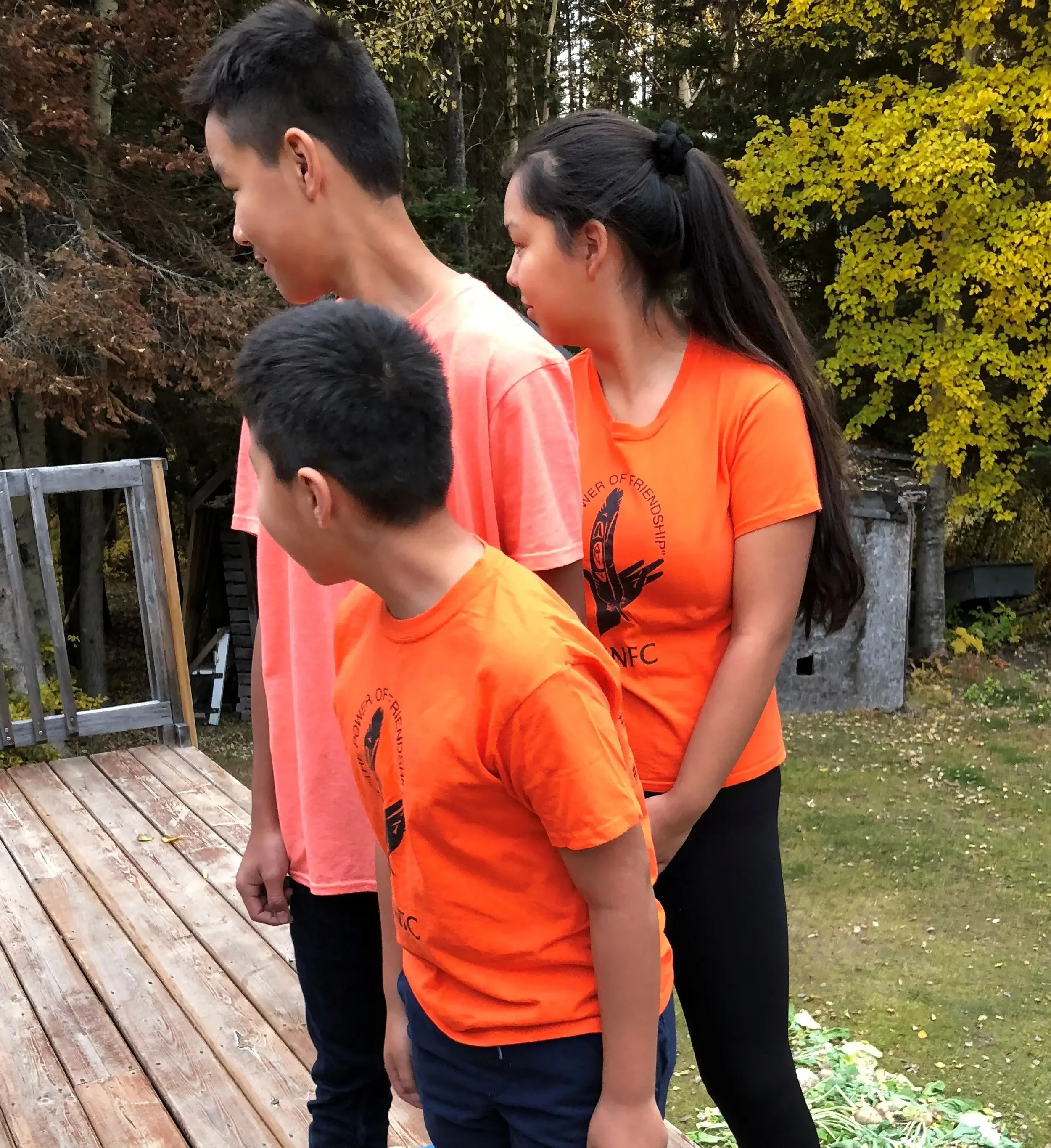 Three children standing outdoors, looking downwards. Two boys in orange shirts with black graphic logos, and a girl in an orange shirt, in front of a wooden deck and trees with yellow and brown leaves.