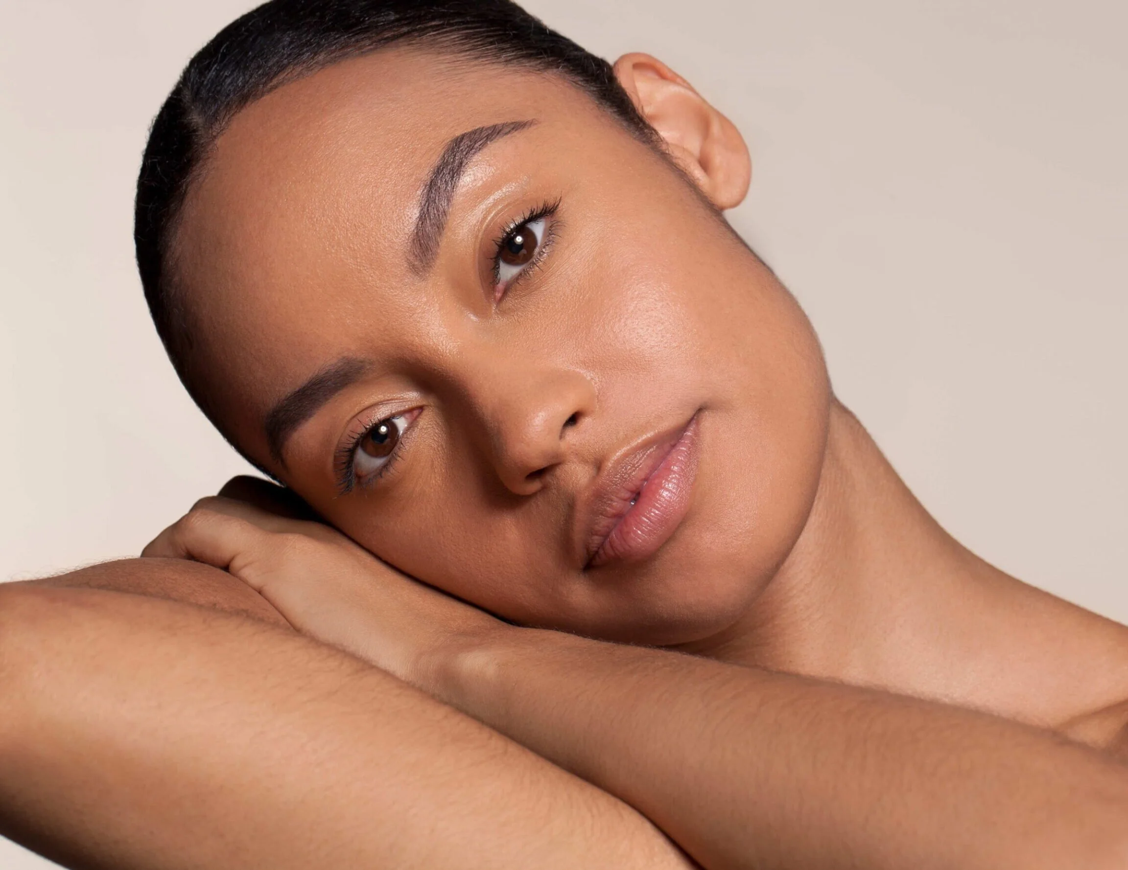 Close-up of a woman with short dark hair, resting her head on her arm, with a serene facial expression, smooth skin, and subtle makeup, against a neutral background.