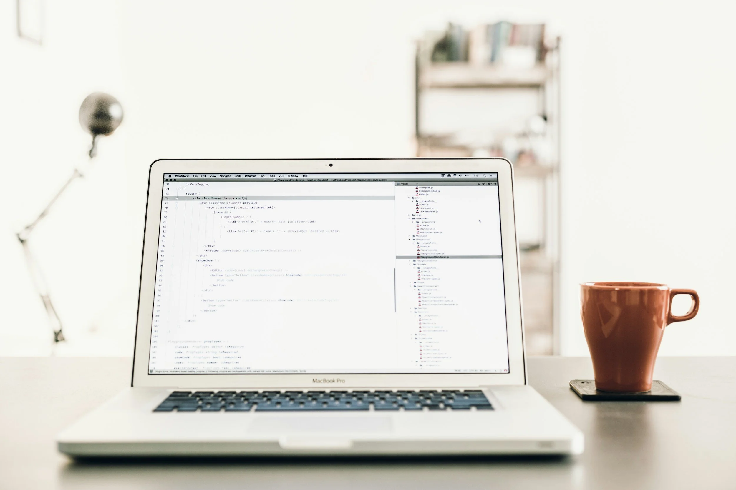 A MacBook Pro laptop open on a white desk, displaying code on the screen. To the right of the laptop, there is a brown ceramic mug on top of a small black notebook or pad. In the background, there is a blurred bookshelf and a metallic object hanging on the wall.