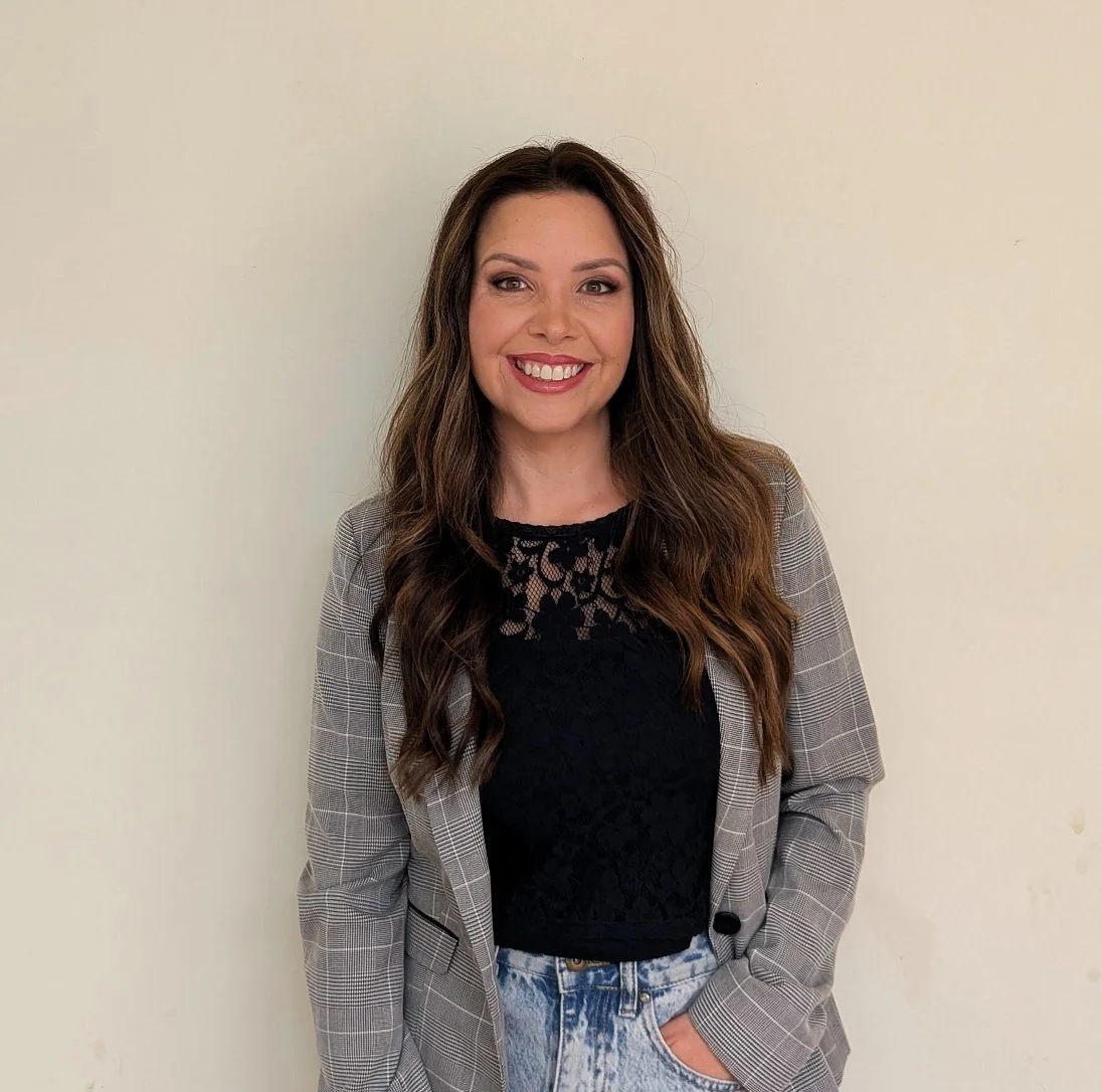 Woman with long wavy brown hair smiling, wearing a black lace top, a plaid blazer, and acid-wash jeans, standing against a plain light-colored wall.