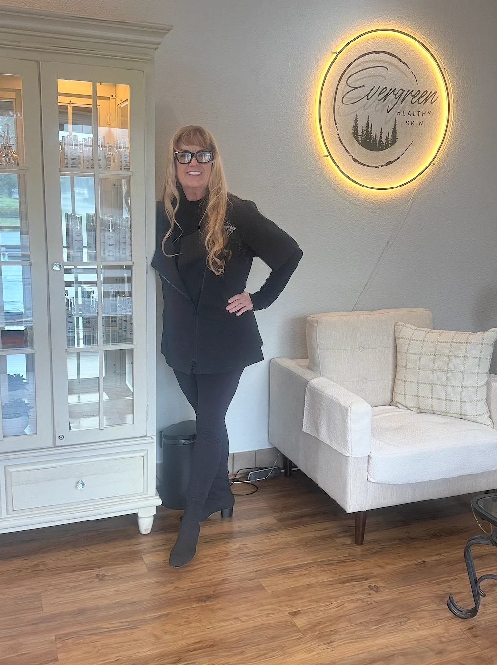 A woman with long blonde hair, glasses, and a black outfit standing in a waiting area of a wellness clinic named Evergreen, with a circular illuminated sign on the wall behind her that says 'Evergreen Healthy Skin.' The area has a cream-colored sofa and wooden flooring.