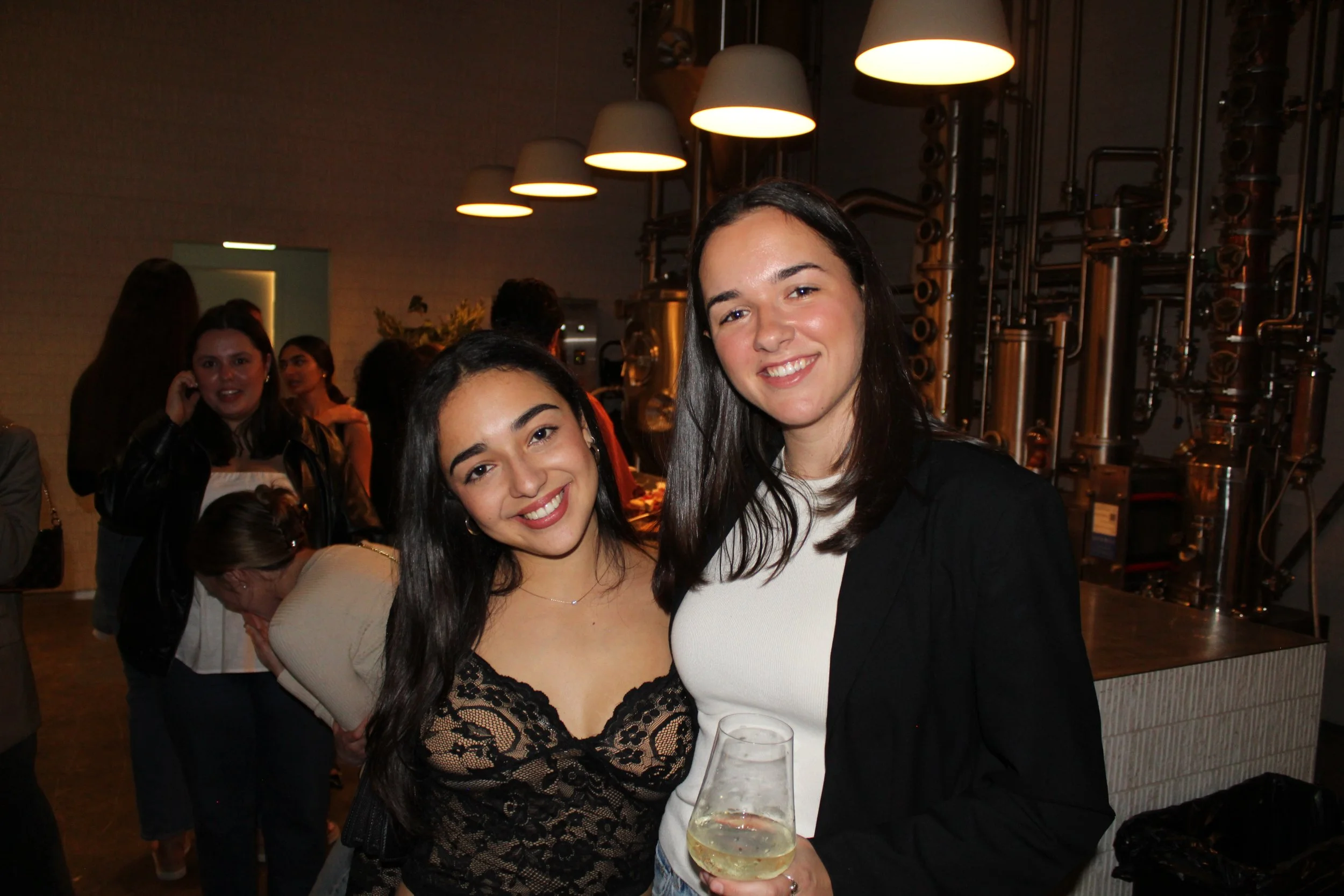 Two women smiling and posing for a photo at a social gathering with dim lighting and industrial decor, one holding a glass of white wine, in an indoor setting.