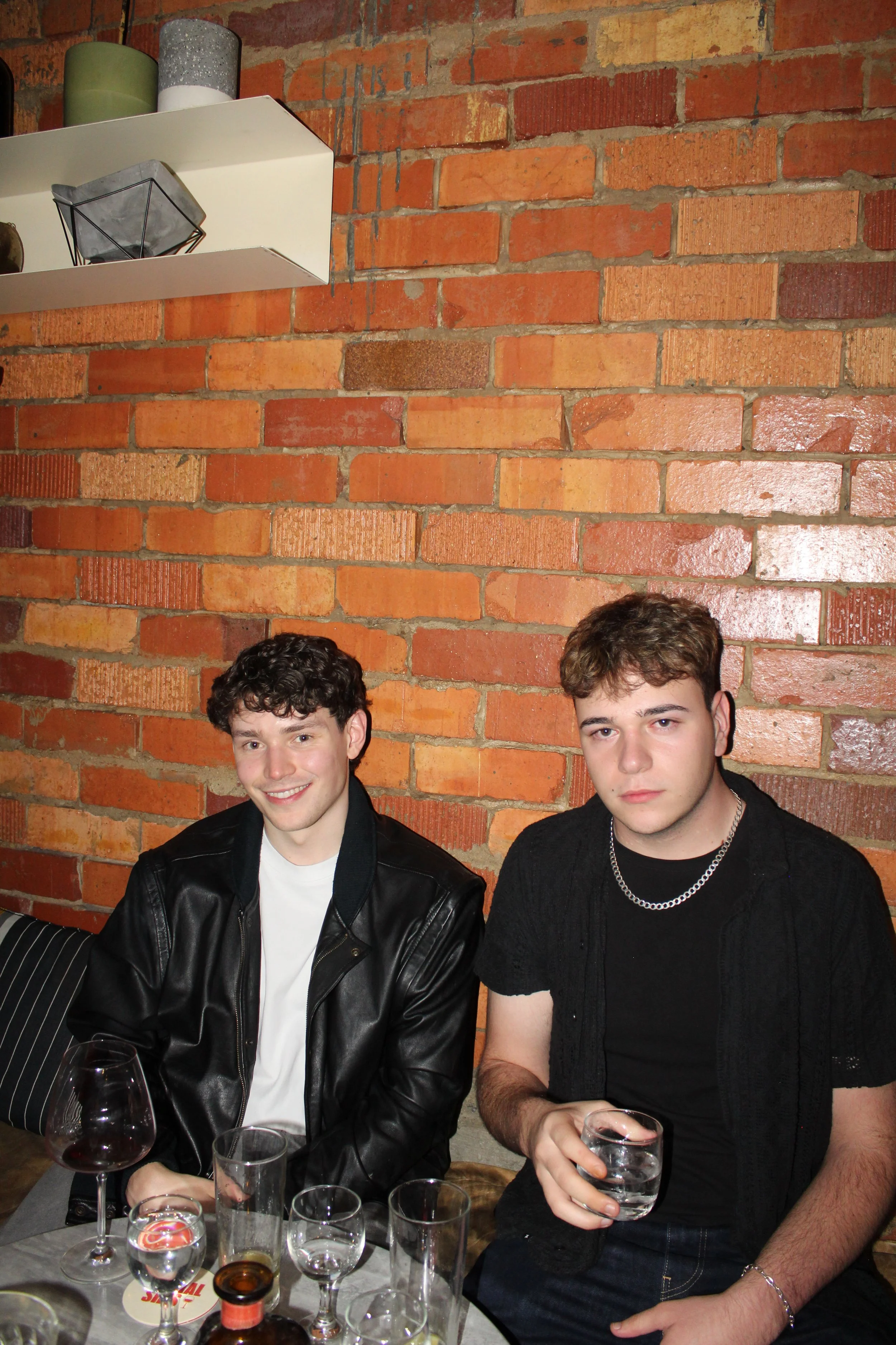 Two young men sitting at a table with drinks in a restaurant or cafe with a brick wall background. One is wearing a black leather jacket and the other a black t-shirt with a silver chain. They are partially visible from the chest up, smiling and look