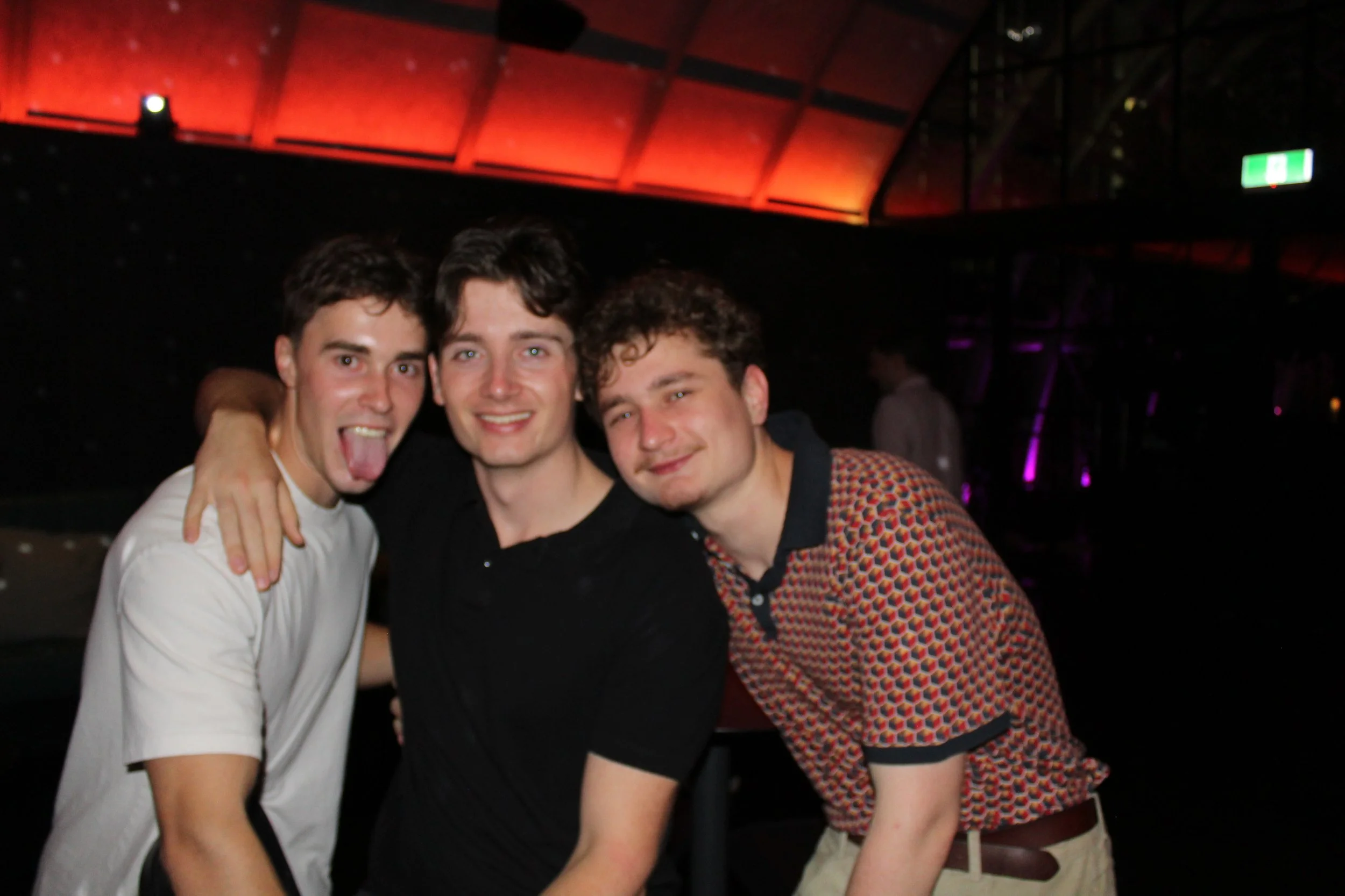 Three young men posing together at a nightclub or party, with colorful lighting and a dark background.