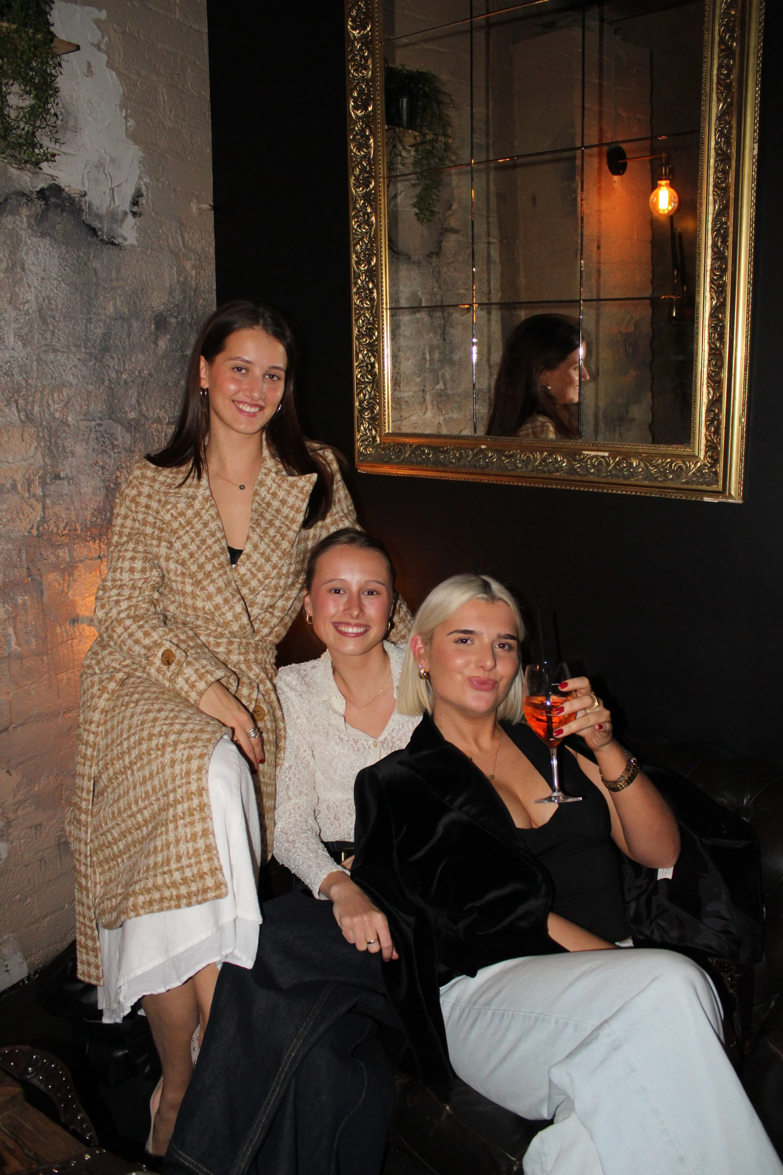 Three women at a nighttime gathering, one holding a glass of rosé wine, sitting and standing in a cozy, dimly lit indoor space with exposed brick walls, decorated with a large ornate mirror.